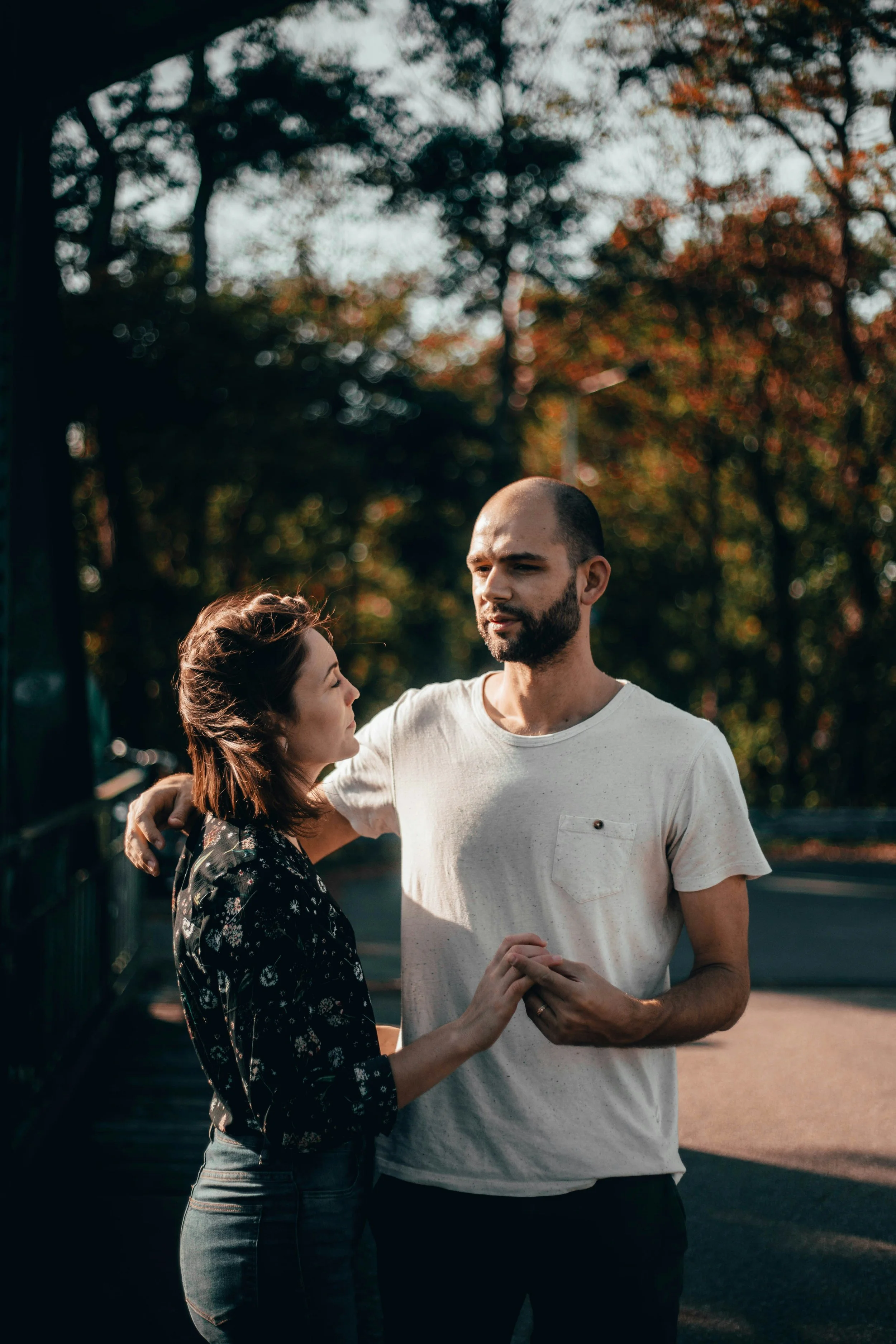 A man and woman are standing closely together outdoors, holding hands, with a background of trees and sky, engaged in a conversation.