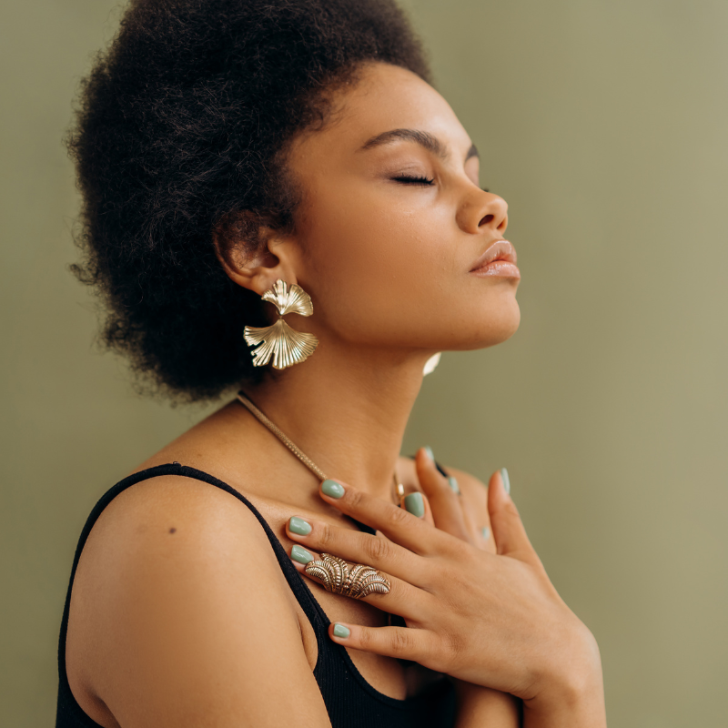A young woman with curly hair and gold earrings, wearing a black top, closes her eyes and touches her chest with her hands, appearing peaceful.