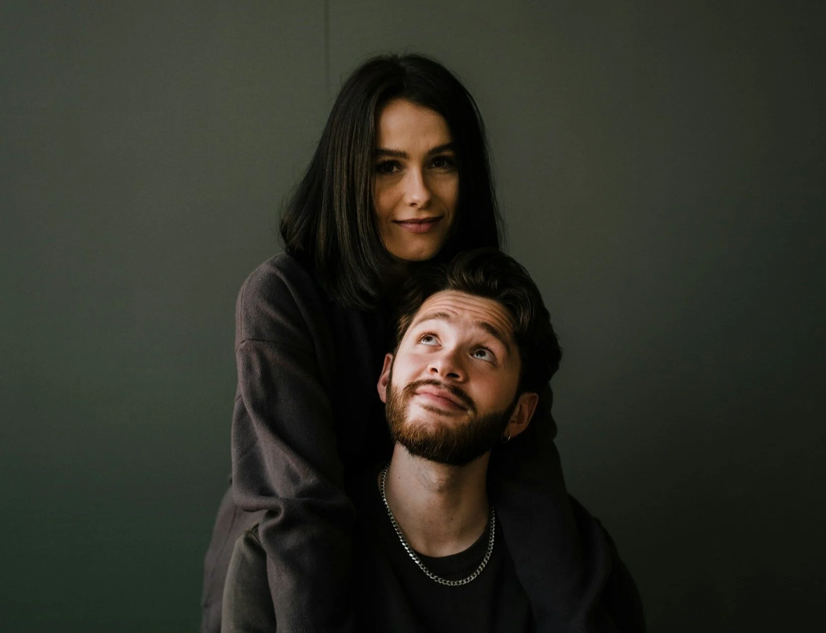Two people, a woman with black hair and a man with a beard, are posing together against a plain dark background. The woman is standing behind the man, with her arms around his shoulders, and they are both looking at the camera.
