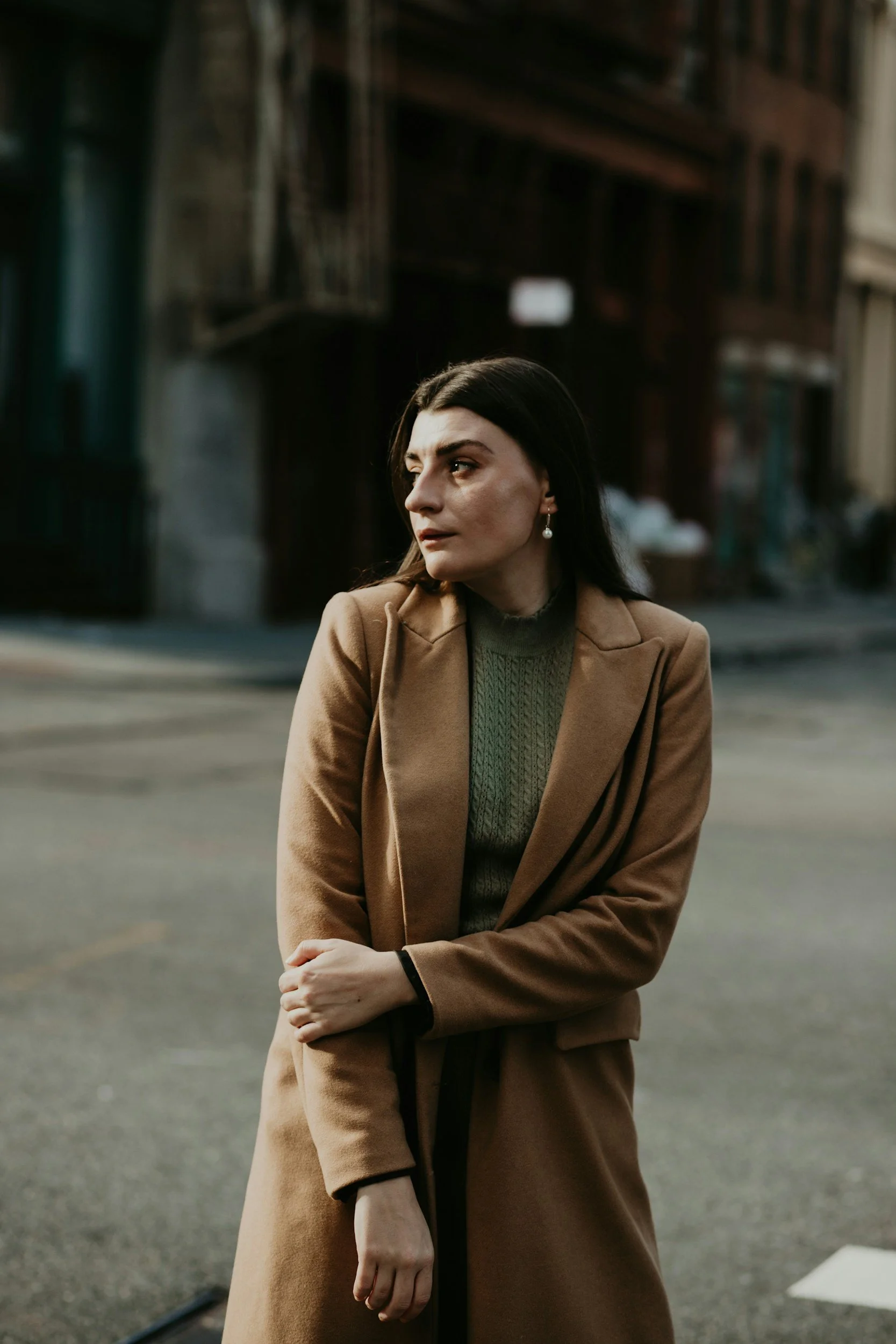 A woman with dark hair wearing earrings and a green sweater, dressed in a brown coat, standing on a city street with buildings in the background.