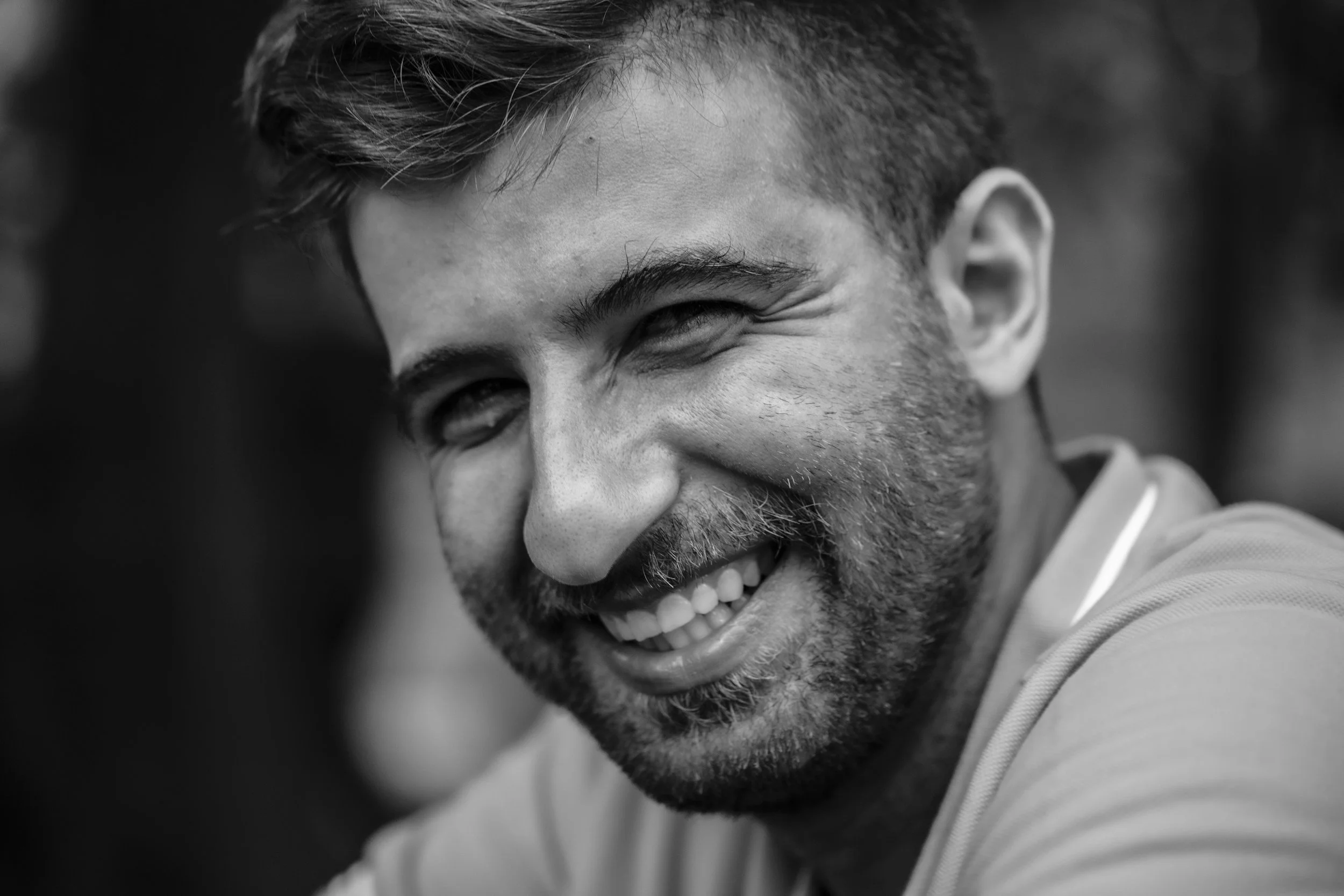 A close-up black and white photo of a smiling man with short hair, a beard, and mustache, showing his teeth and crinkling his eyes.