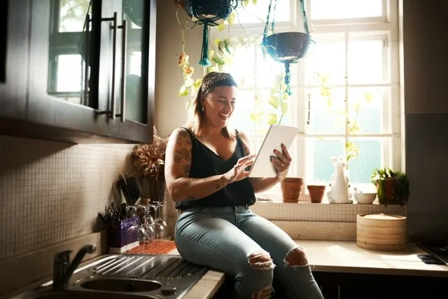 A woman with tattoos, wearing a black tank top and ripped jeans, sitting on a kitchen counter, smiling while using a tablet. The kitchen has hanging plants, sunlight coming through a window, and various kitchen items around.