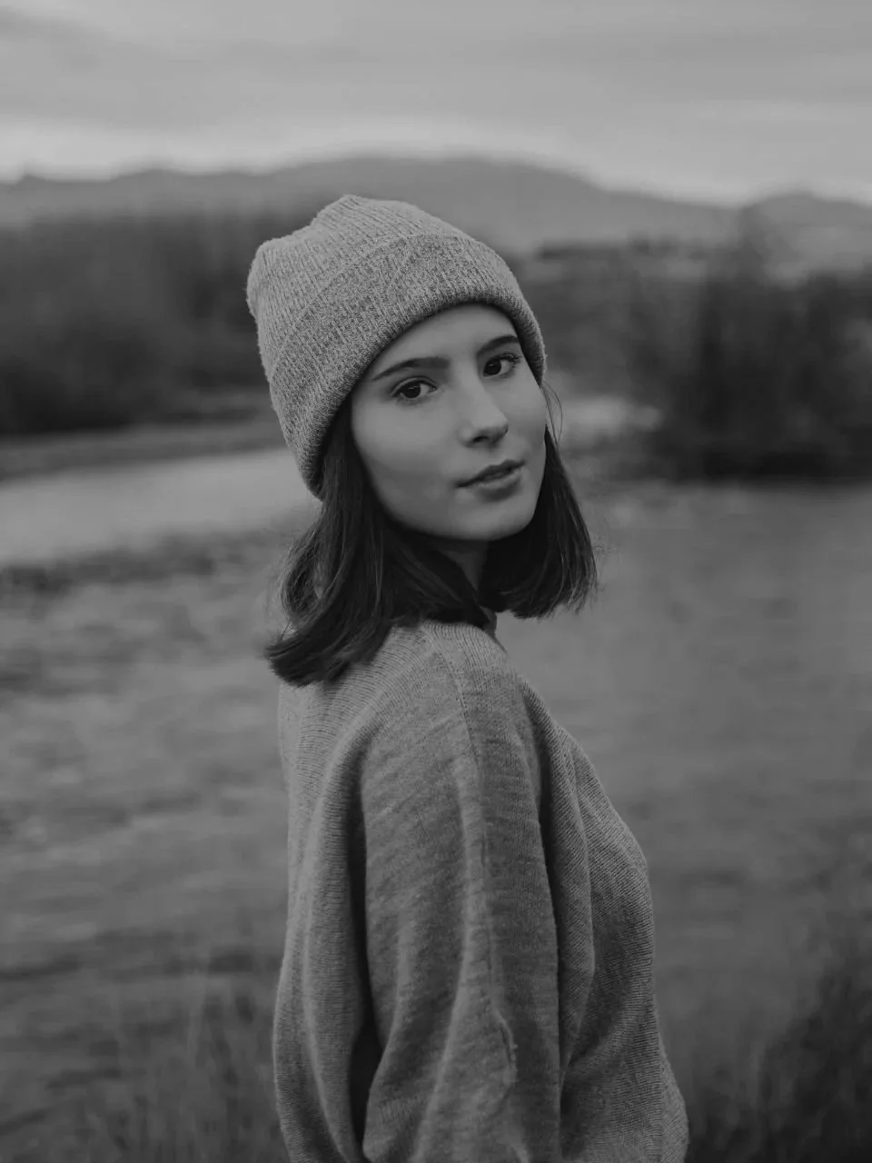 Black and white portrait of a young woman wearing a beanie and a sweater outdoors with a blurred landscape of trees and hills in the background.