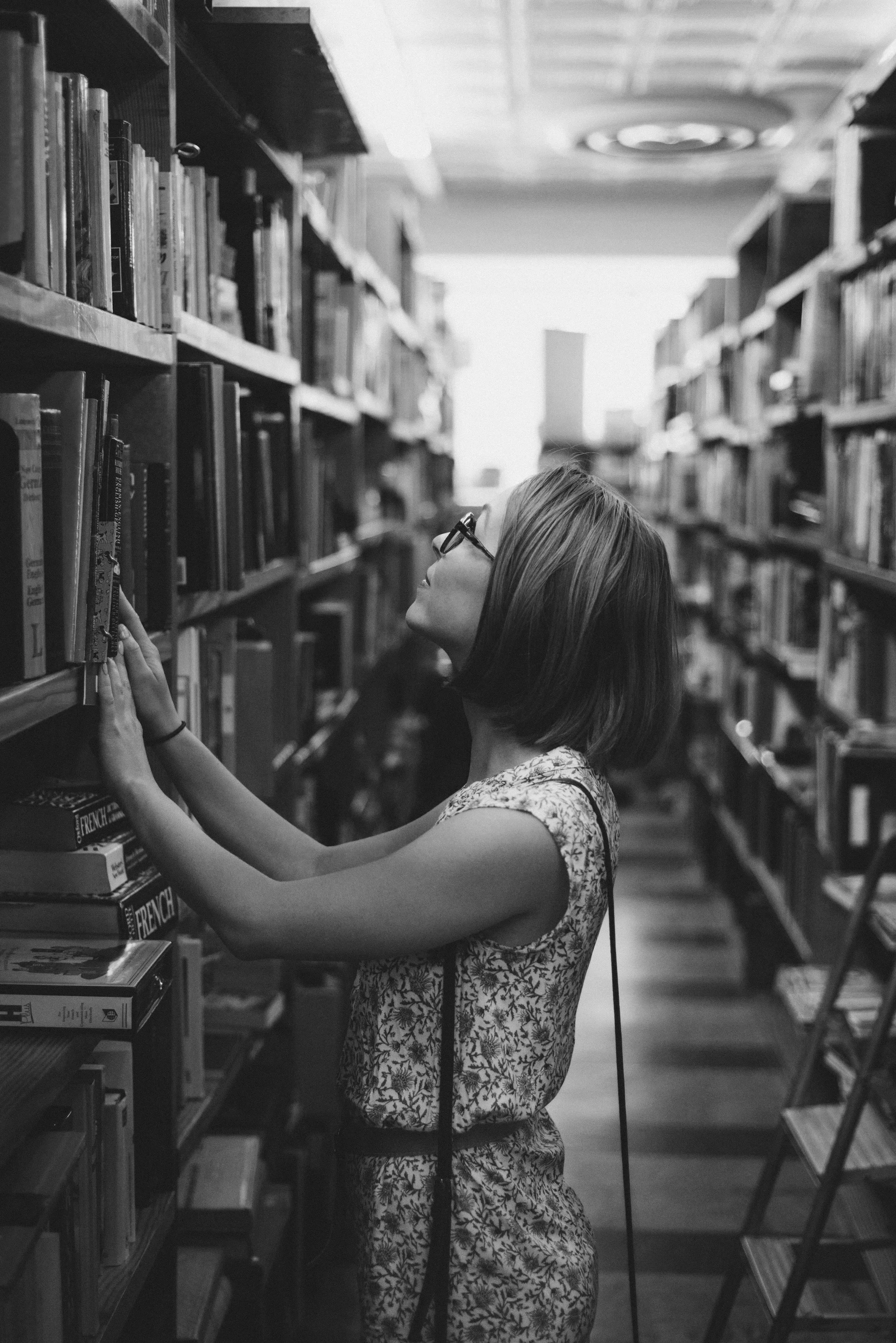 Woman in glasses reaching for a book on a library shelf.