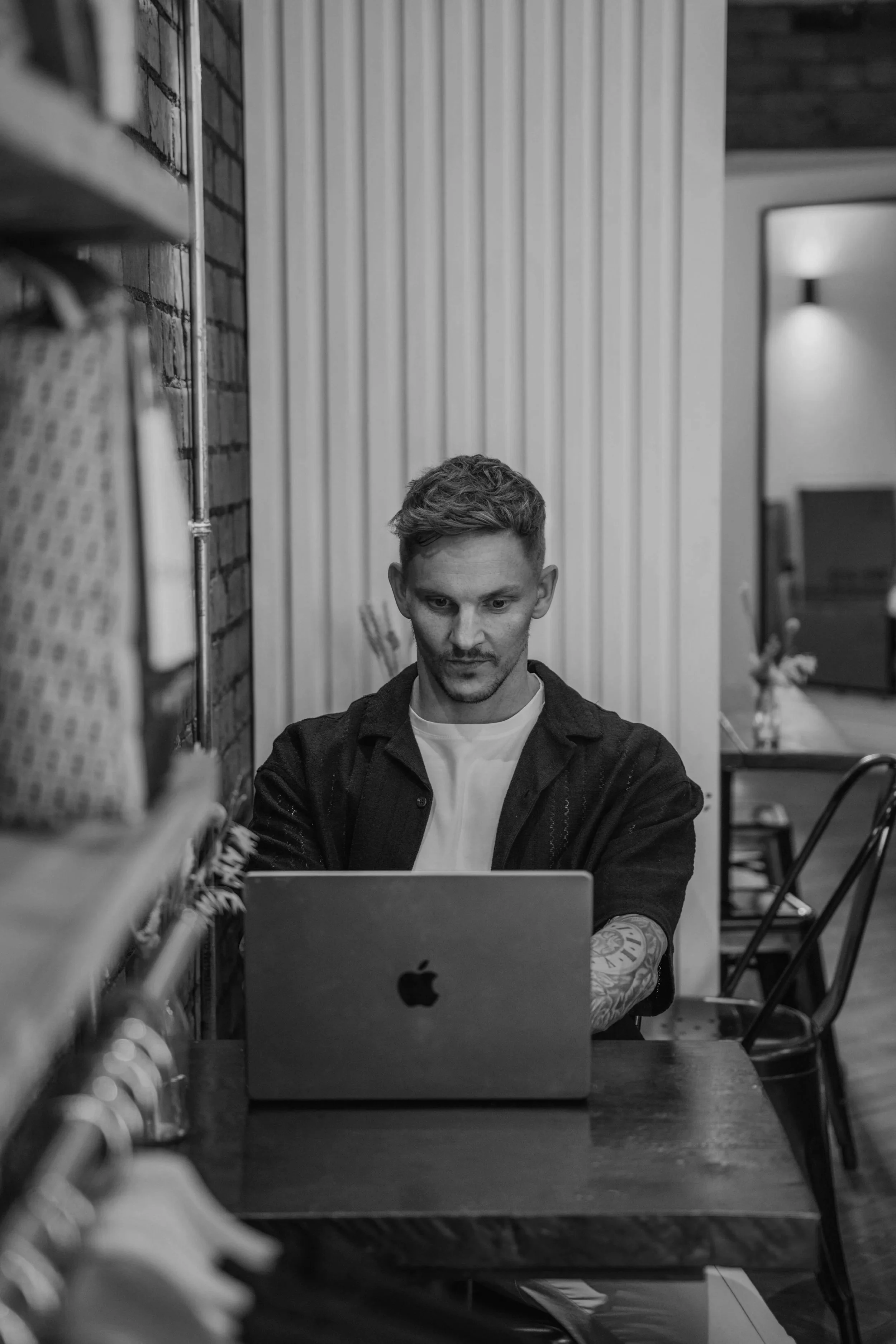 A young man with short, styled hair and tattoos on his arms working on a laptop at a wooden table in a modern, industrial-style space.