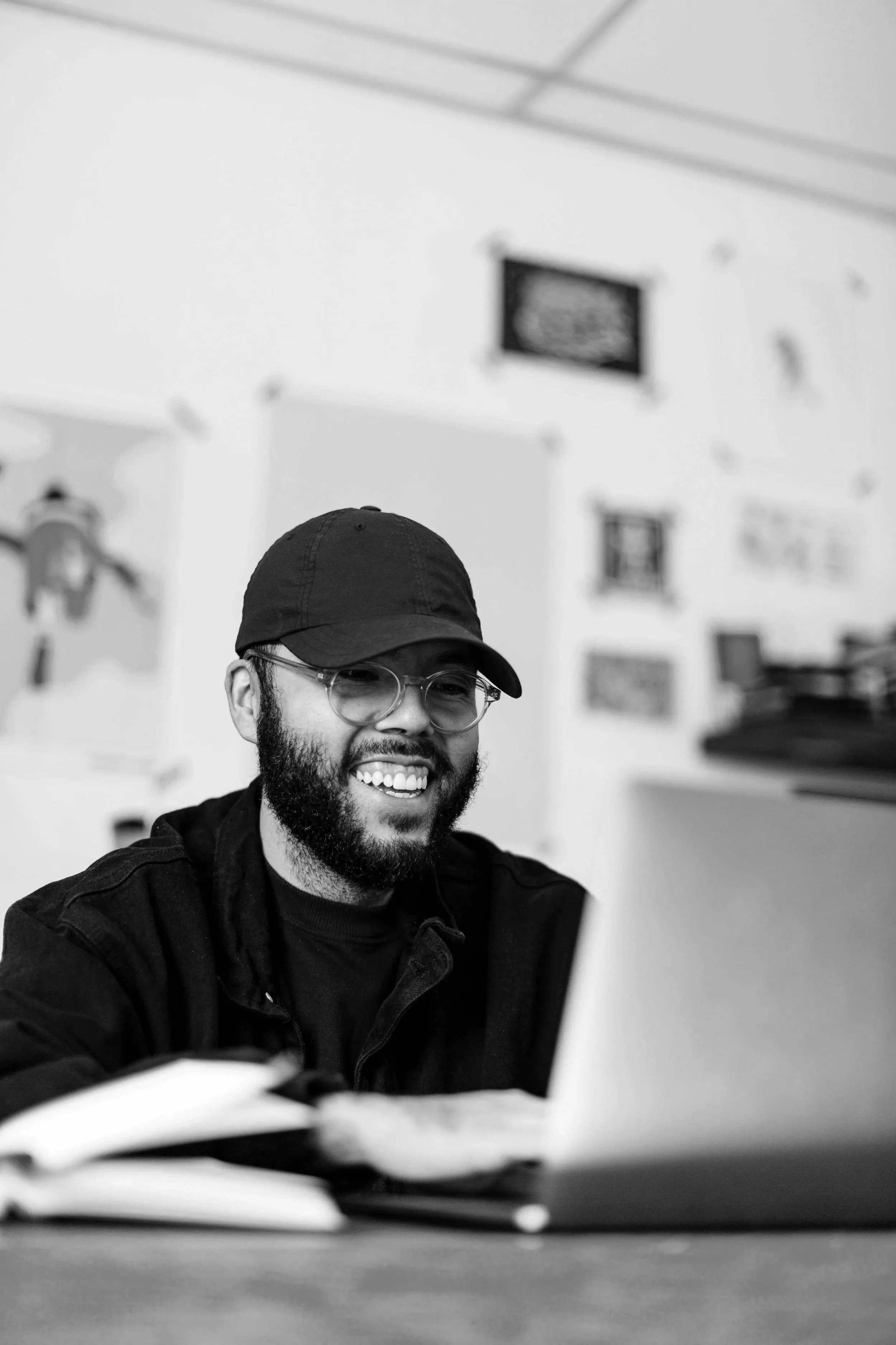 A smiling man with glasses, a beard, and a black cap, sitting at a table with papers or notebooks, in a room with framed pictures on the wall.