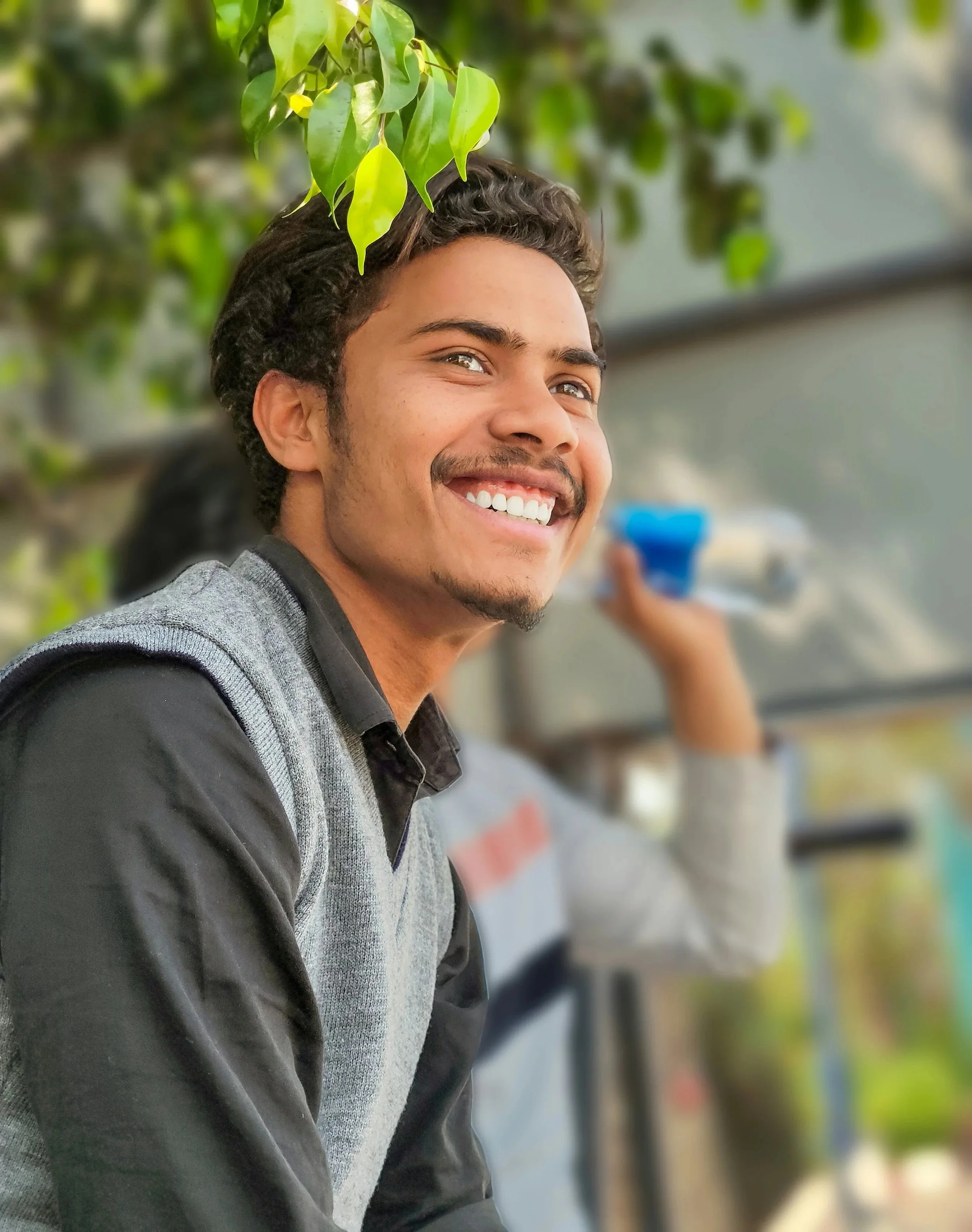 Close-up of a smiling young man with curly hair and a light beard, wearing a gray and black jacket, standing outdoors with a blurred background of green leaves and a structure.