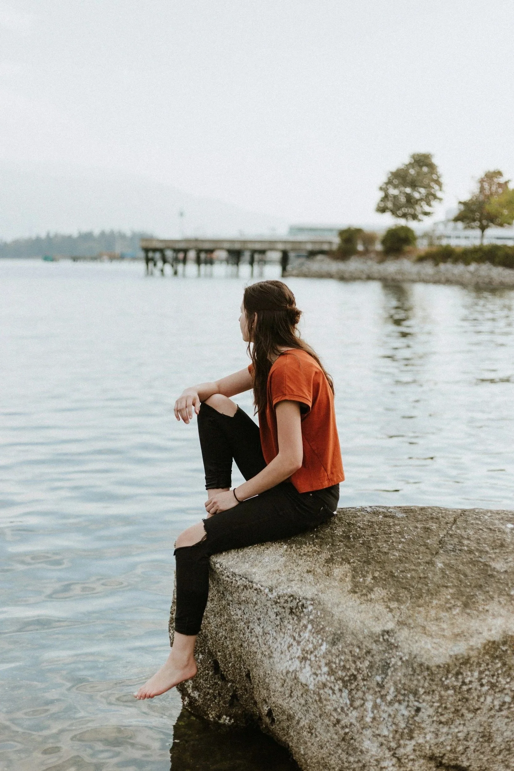 A woman sitting on a rock by the water with her legs dangling into the water, near a pier, trees, and a building in the background, on a cloudy day.