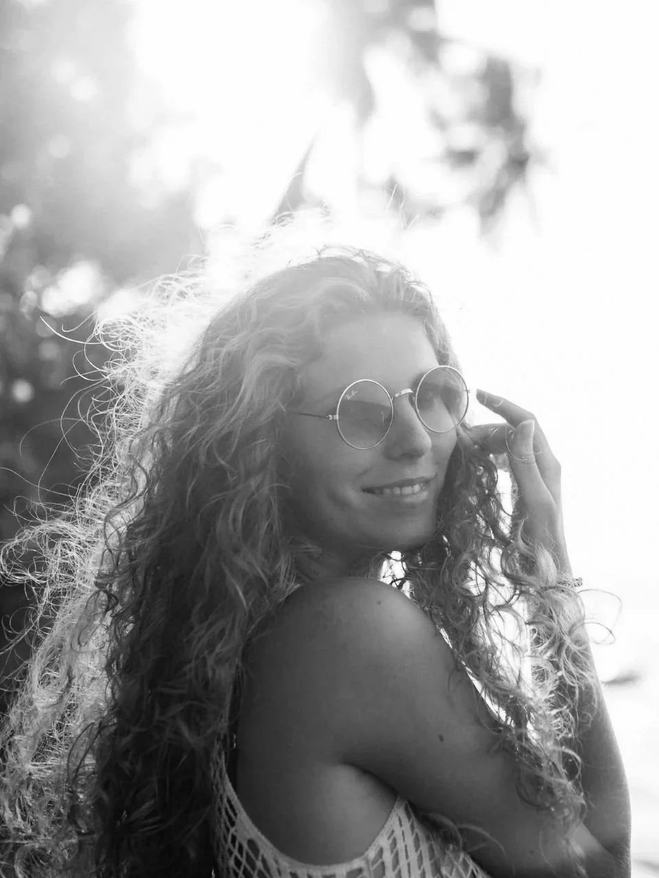 A woman with curly hair and round sunglasses smiling, with a person waving in the background at sunset or sunrise.