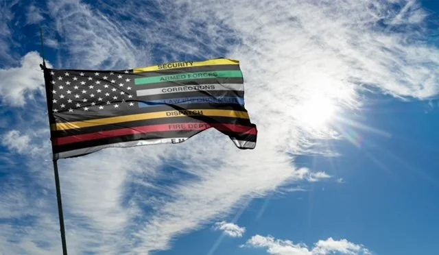 Flag with black and white American stars and stripes, overlaid with colored stripes representing different military or emergency services, flying in a partly cloudy sky.