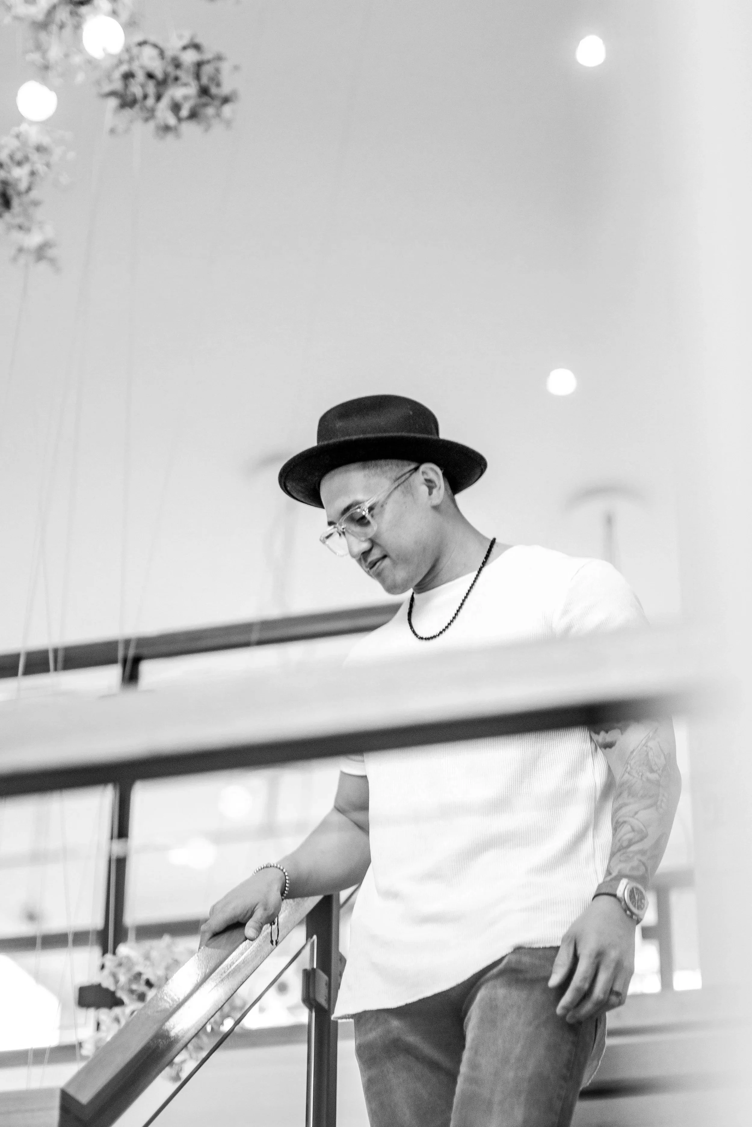 A young man wearing glasses, a fedora, and casual clothing, leaning on a railing in a modern indoor space with bright lighting and hanging light fixtures.