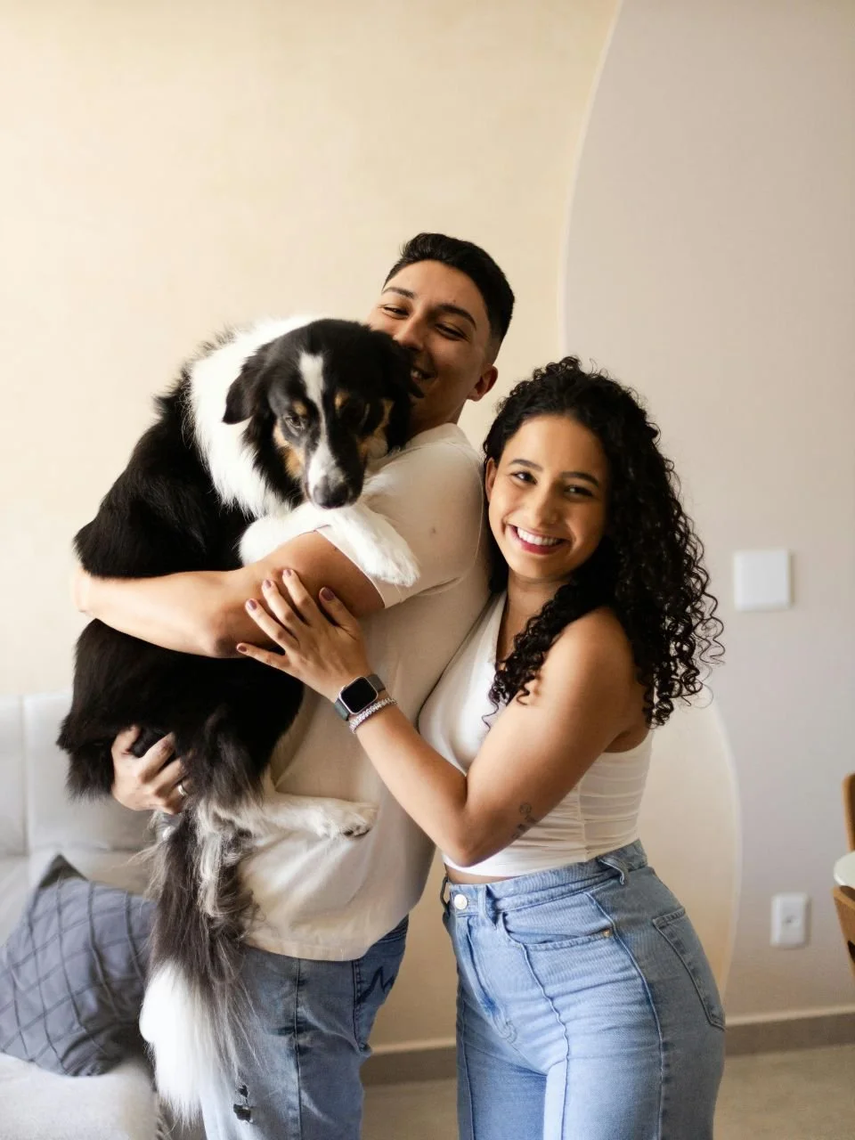A smiling couple with the man holding a black and white dog, standing in a cozy room with neutral-colored walls.