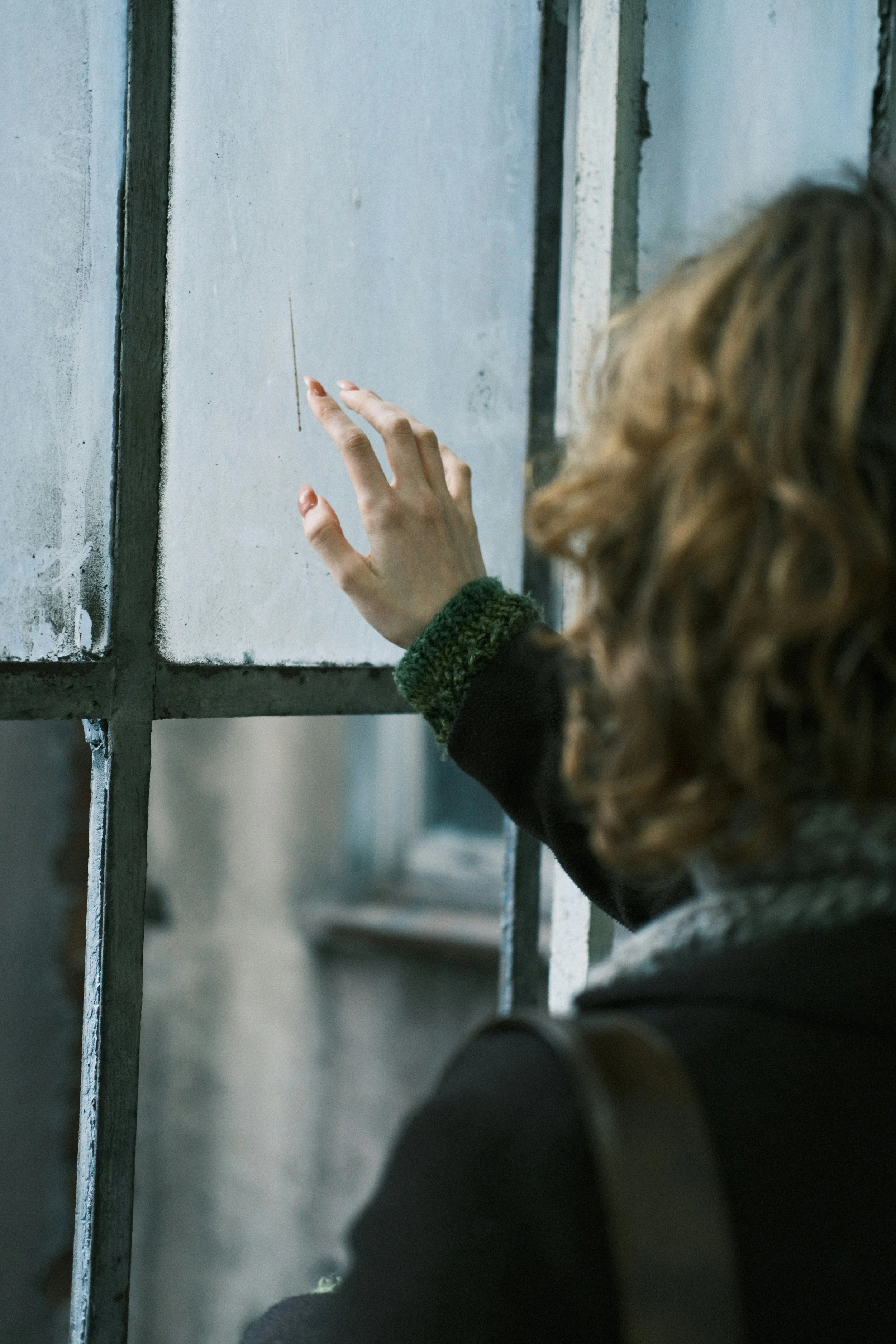 A person with curly hair touching a glass window or barrier with a finger, the person wearing a dark jacket with a green cuff, and metal bars or railing in the foreground.
