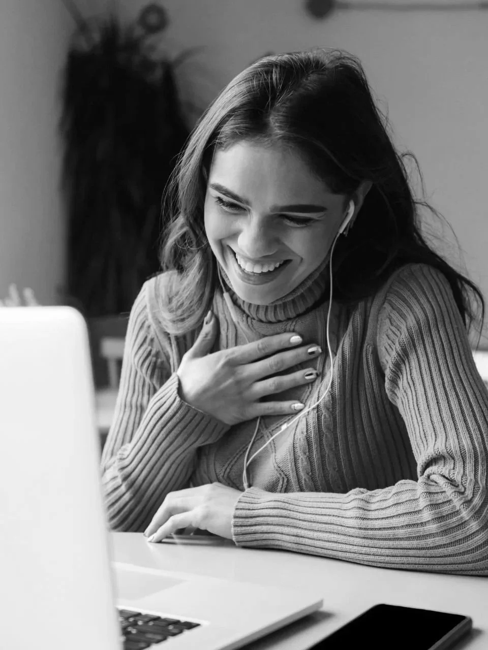 A woman with long hair wearing a turtleneck sweater, smiling and looking at a laptop, with earphones plugged in, sitting at a desk.