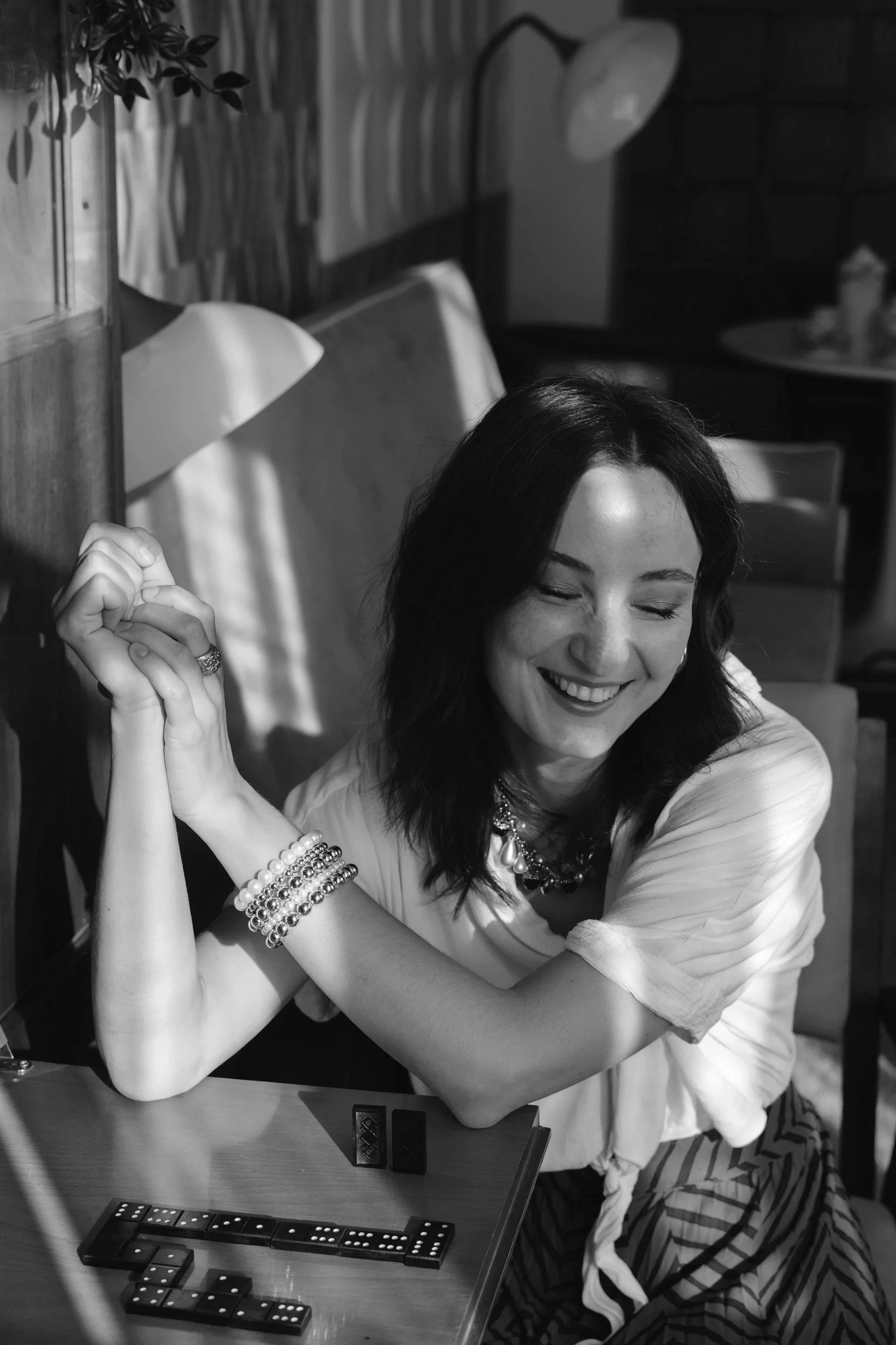 A woman with dark hair smiling with her eyes closed, sitting at a table with domino game pieces arranged in front of her. She has multiple bracelets on her wrist and is wearing a light-colored top. The background shows a wooden wall and some furniture.