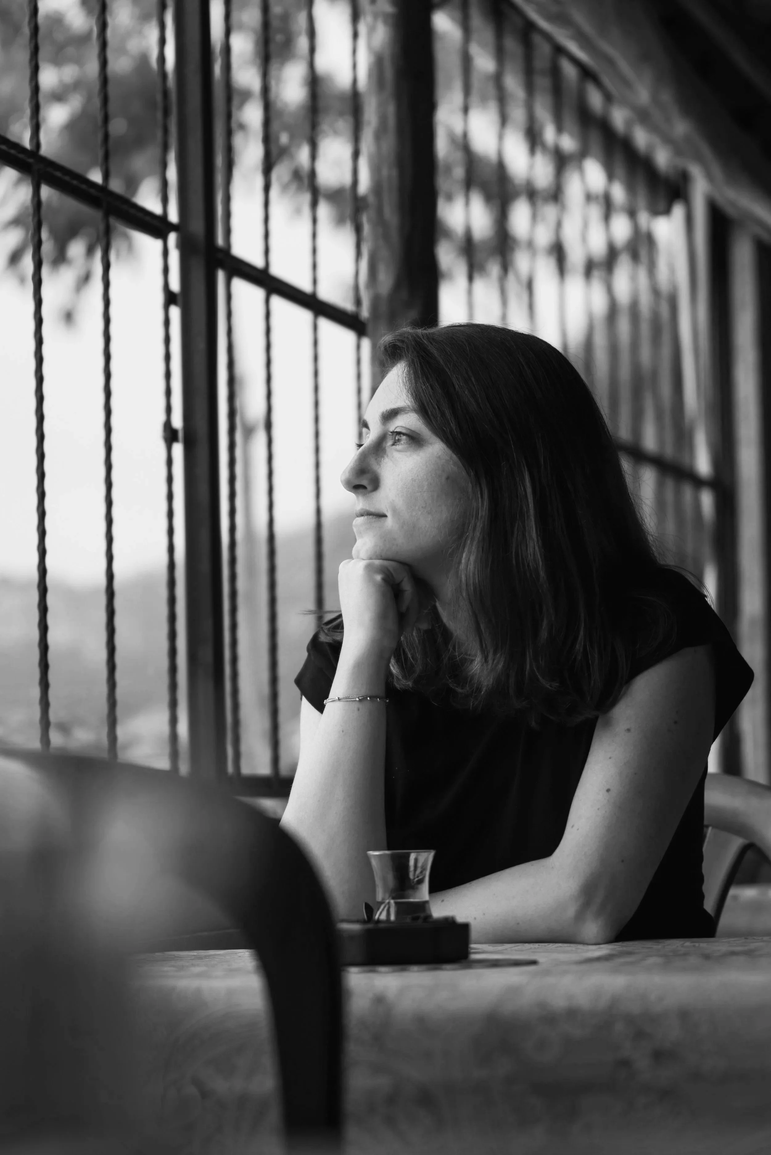 Black and white photo of a woman with dark hair, sitting at a table, resting her chin on her hand, gazing thoughtfully to the side. A glass with a beverage is on the table. The background features a large window with bars, and blurred outdoor scenery.
