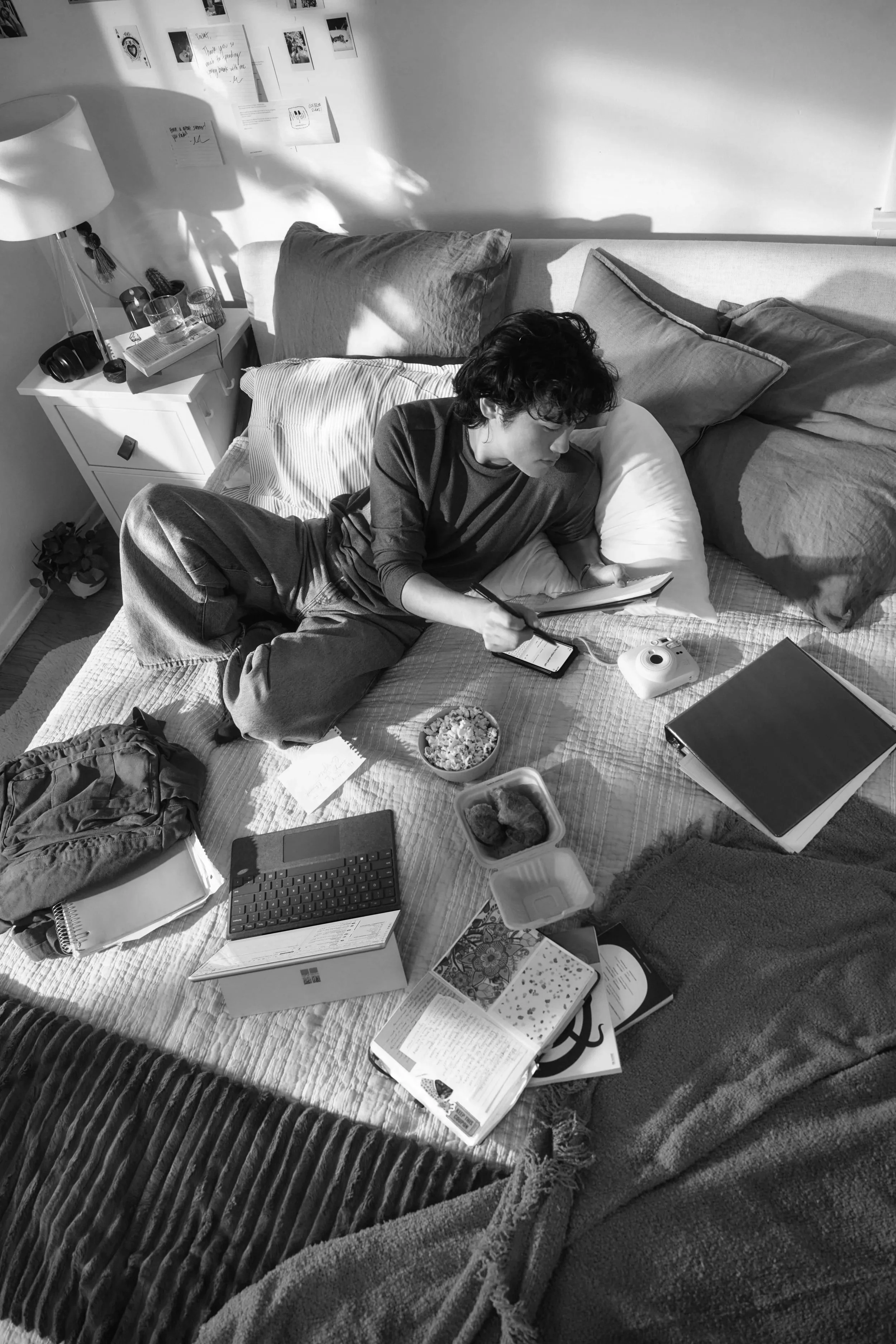 A person lying on a bed surrounded by books, notebooks, a keyboard, a phone, a bowl of popcorn, and various other items, with sunlight casting shadows on the wall.
