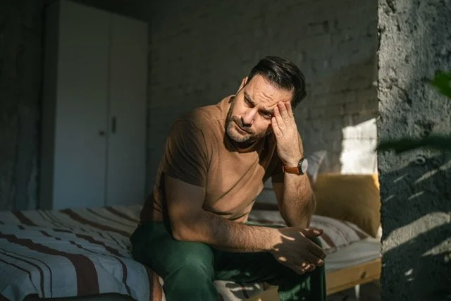 Man sitting on a bed holding his head, appearing distressed or in pain, in a room with brick wall and cabinet in background.