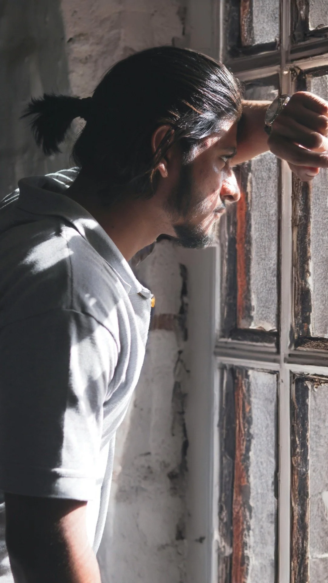 A man with dark hair in a ponytail leaning against a brick wall, looking out a window with sunlight illuminating part of his face.
