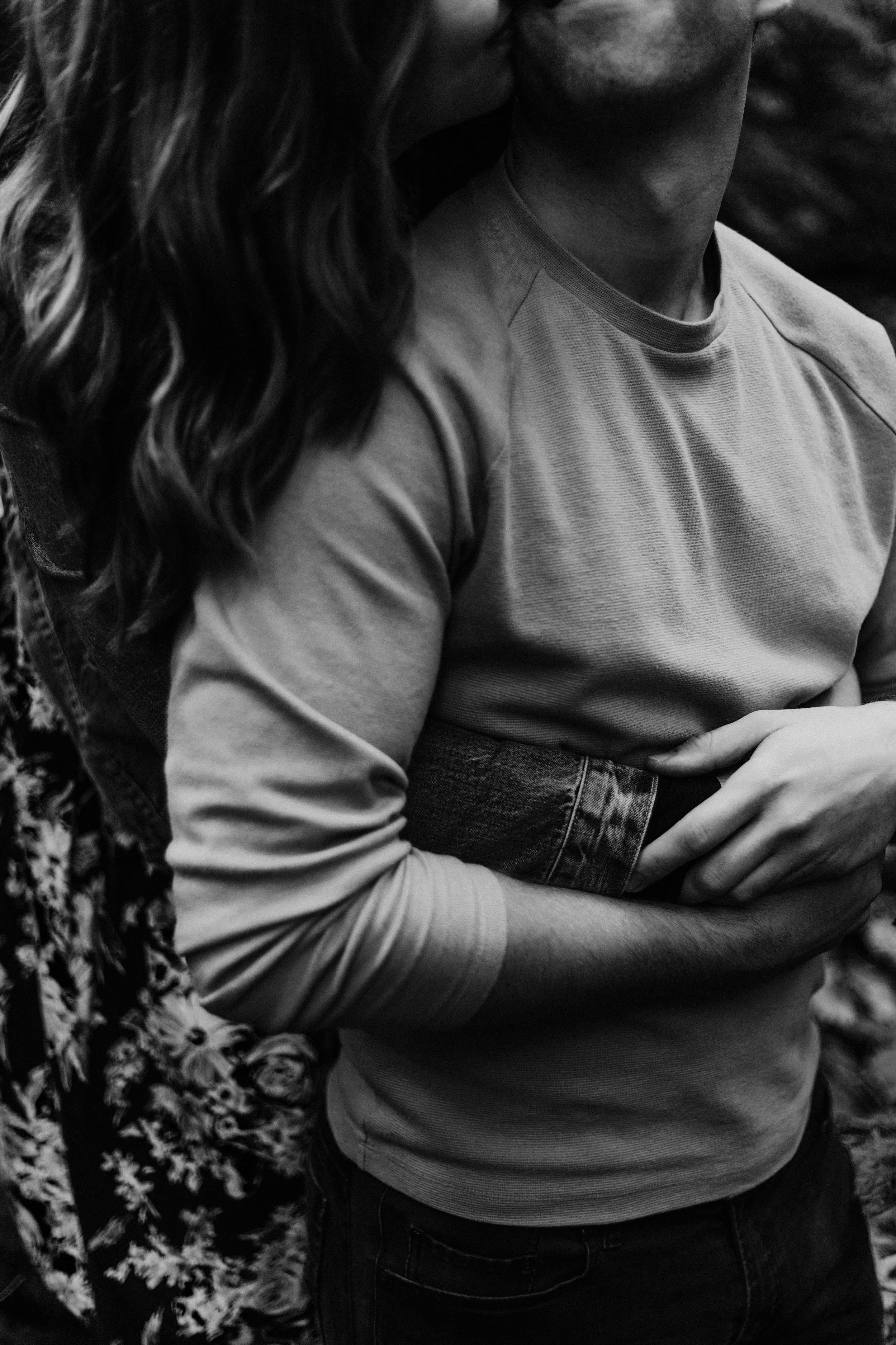 Close-up of two people, one with long, curly hair, hugging each other. The focus is on their arms, torsos, and part of the necks, in black and white.