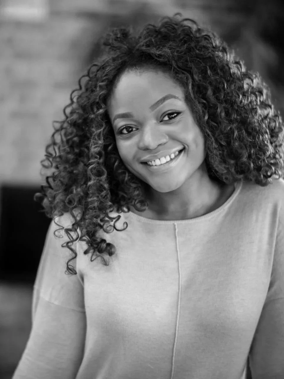 A smiling woman with curly hair wearing a light-colored top, looking at the camera, in black and white.