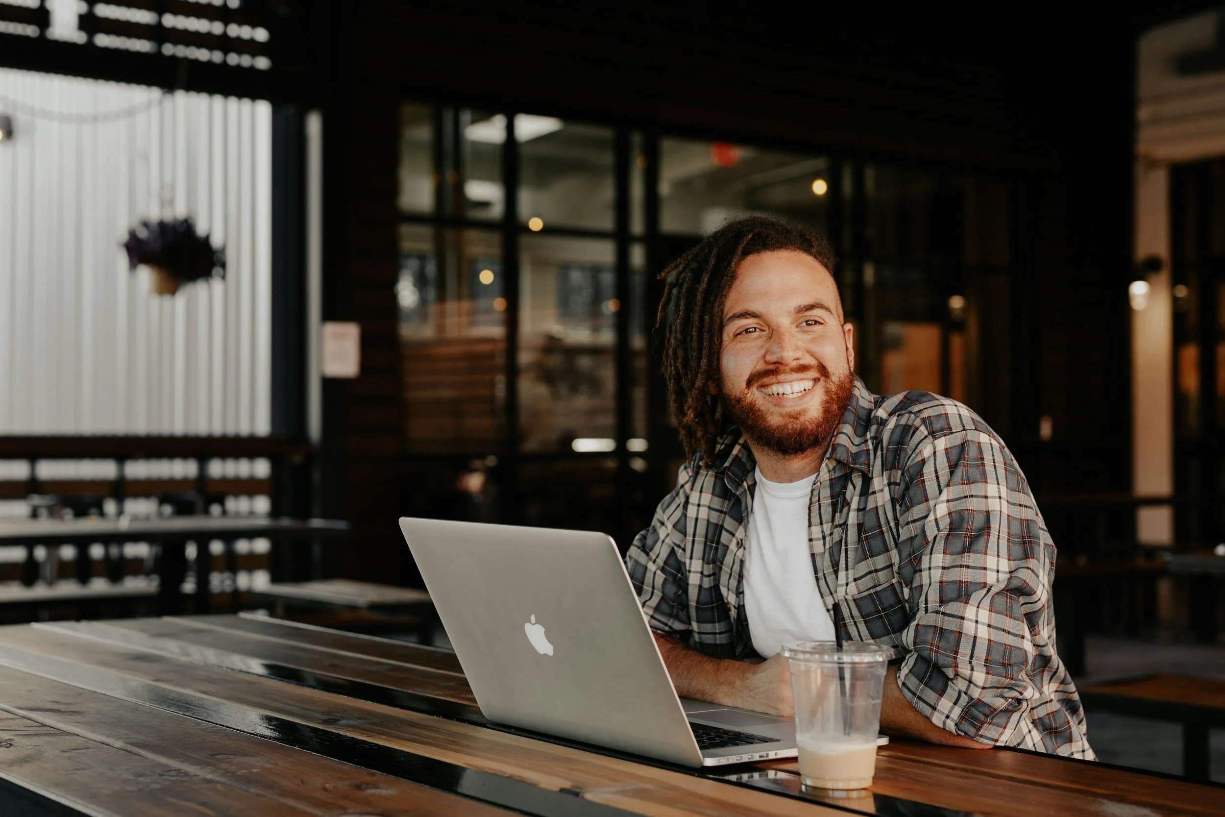 A smiling man with dreadlocks and a beard working on a MacBook laptop in a coffee shop, with an iced coffee on the table and a modern, dimly lit interior in the background.
