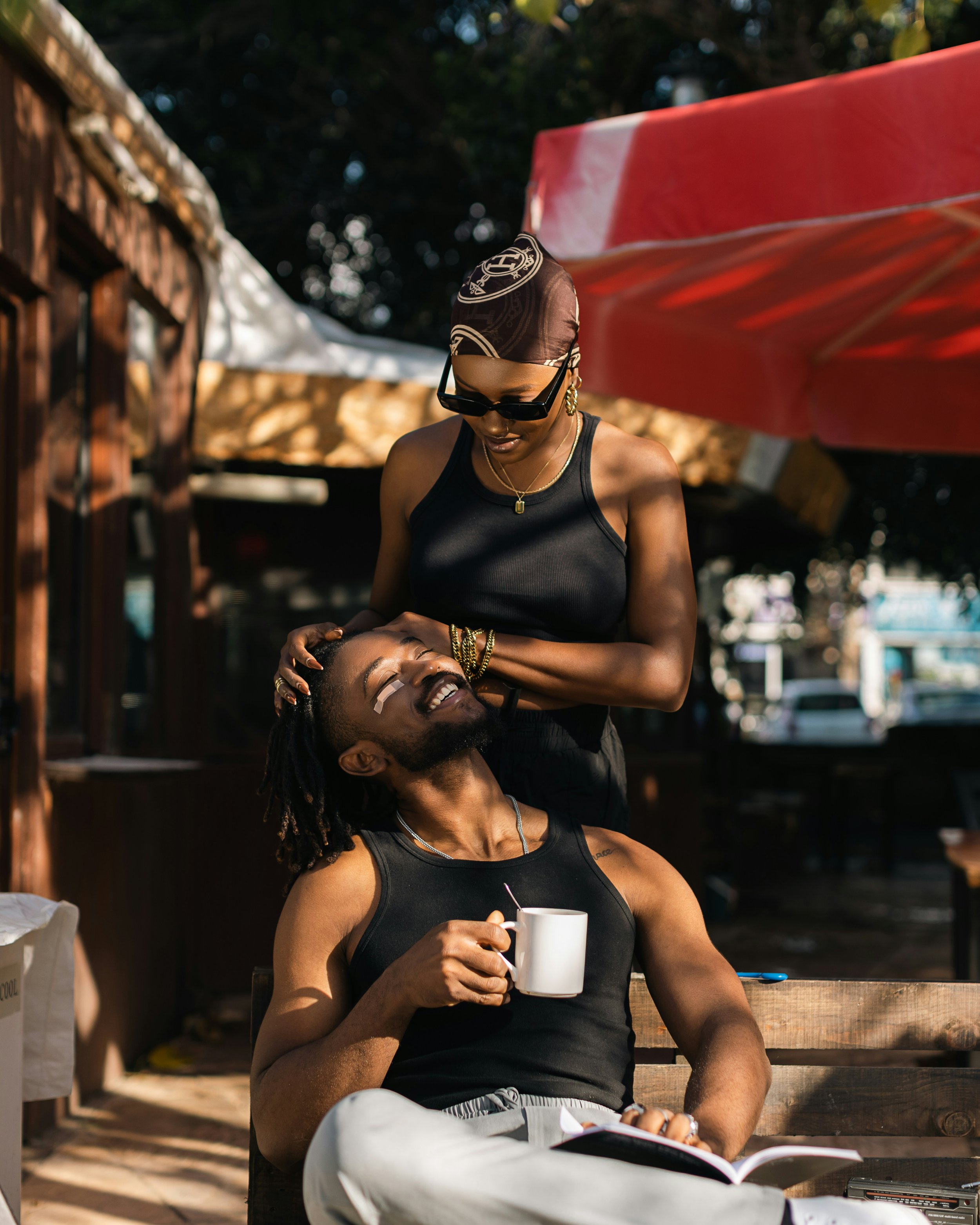 A woman with a headscarf and sunglasses giving a massage to a man with dreadlocks and a beard, who is holding a mug and sitting on a bench outdoors.
