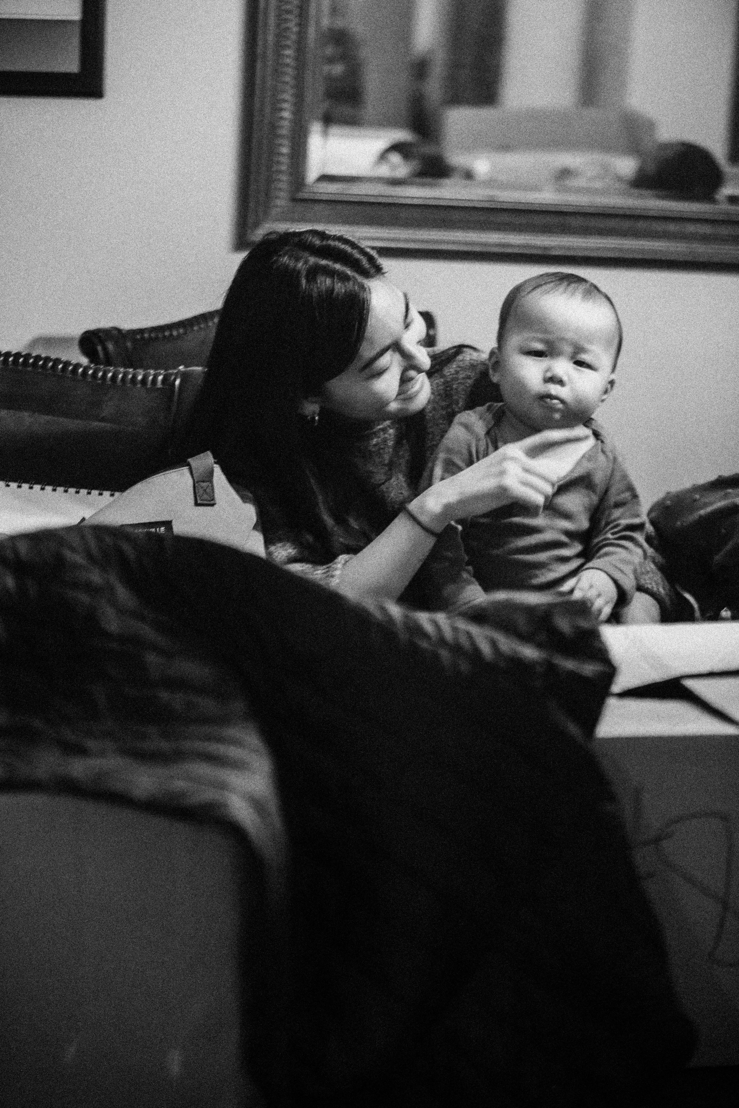 A woman with dark hair holding a young child in a cozy indoor setting, with a large mirror and framed picture on the wall behind them.