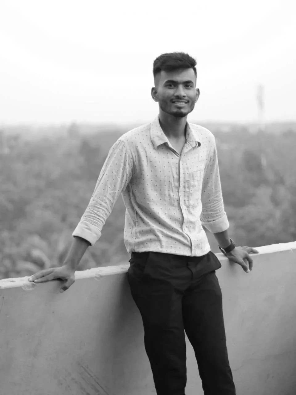 A young man leaning on a railing outdoors, smiling, wearing a button-up shirt and dark pants, with a blurred background.