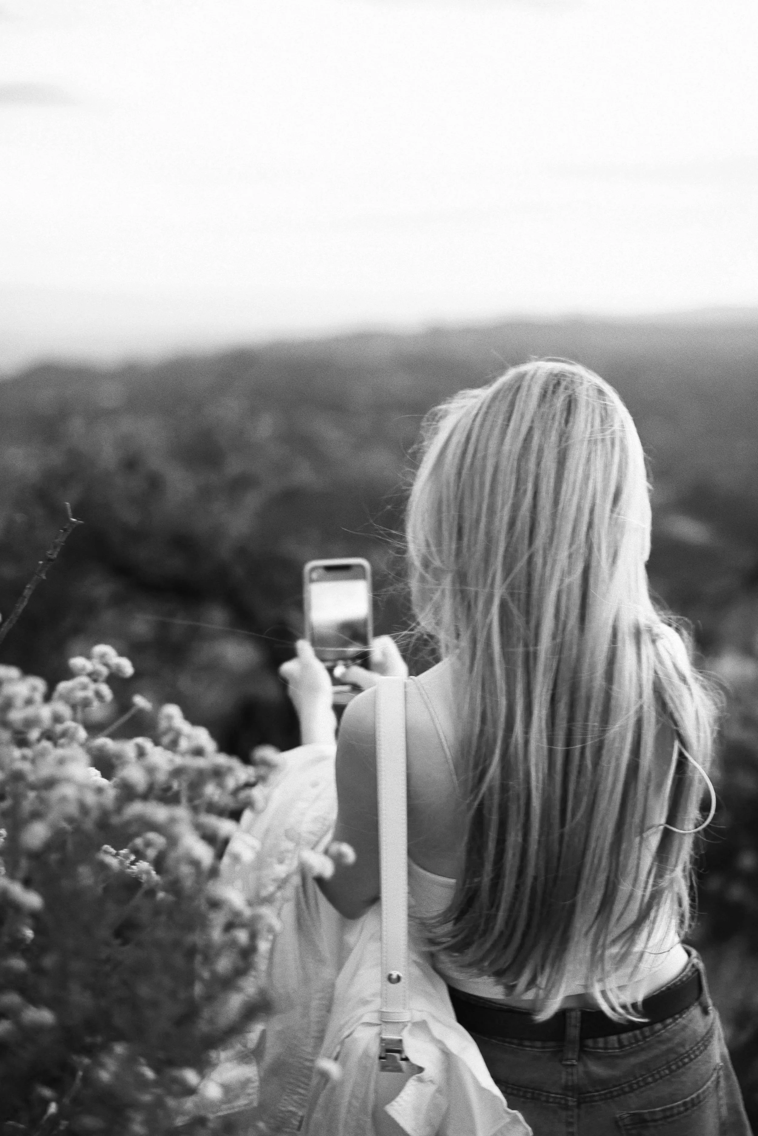 A woman with long hair taking a selfie outdoors, with a landscape of hills or mountains in the background. She is wearing a light-colored jacket and jeans.