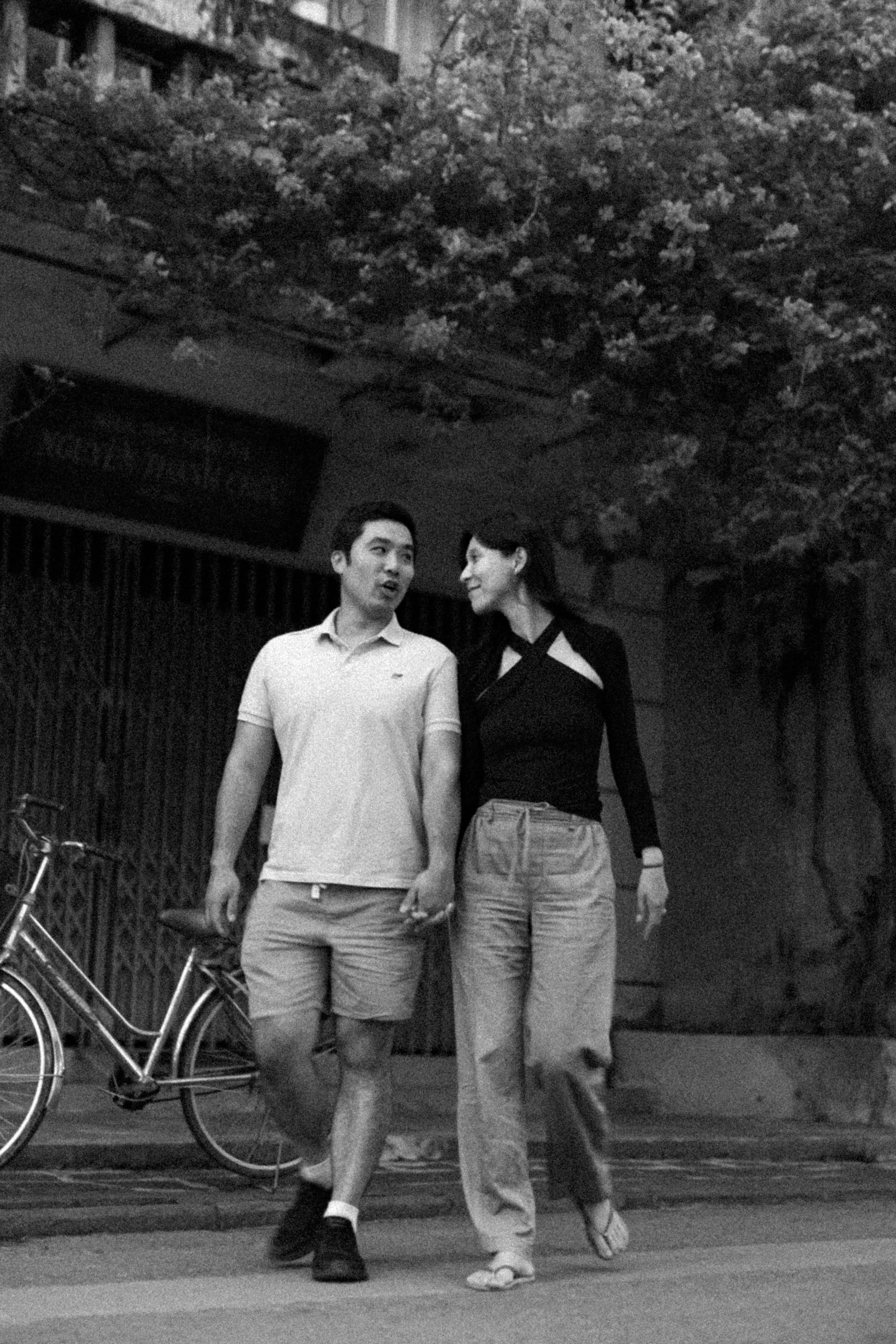 A black and white photo of a young man and woman strolling together in an outdoor setting, with bicycles and a wooden fence in the background.