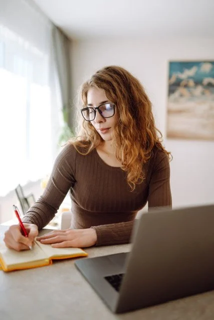 Woman with curly red hair and glasses taking notes at a desk with a laptop in a bright room.