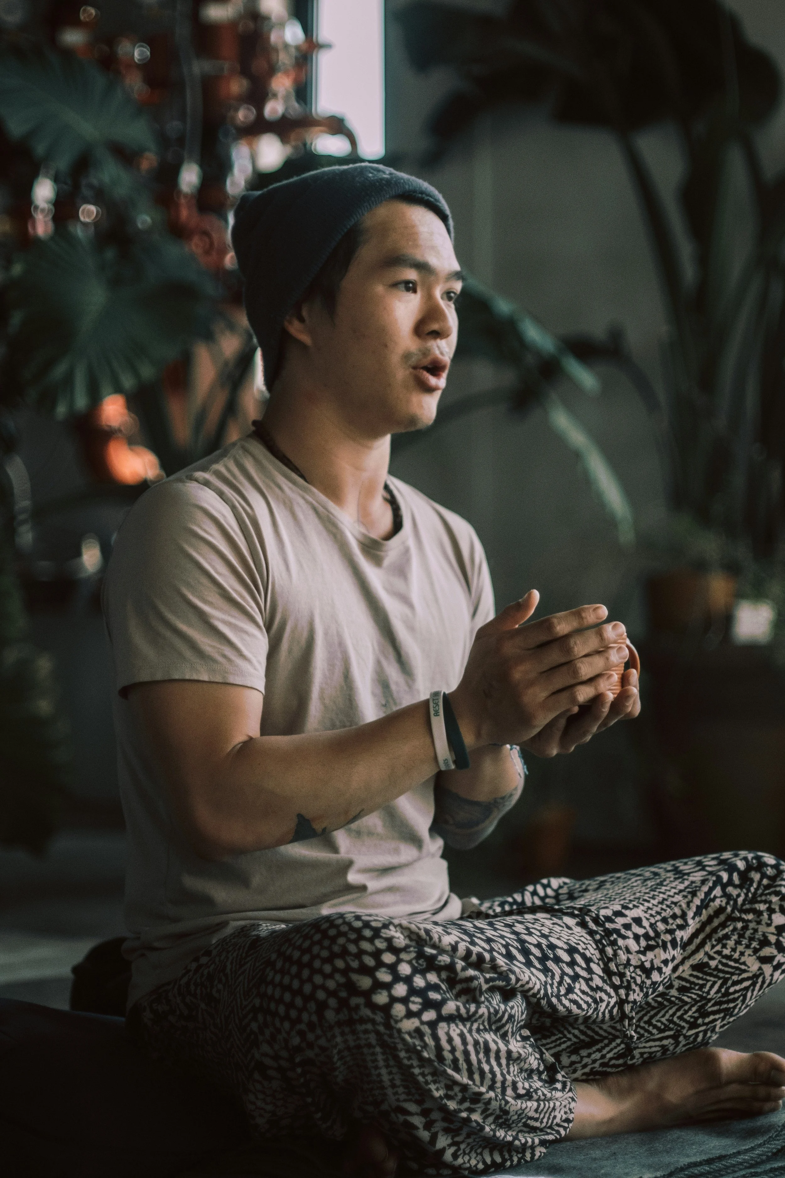 A person sitting cross-legged on a yoga mat, wearing patterned pants, a beige t-shirt, and a beanie, practicing meditation indoors with plants in the background.
