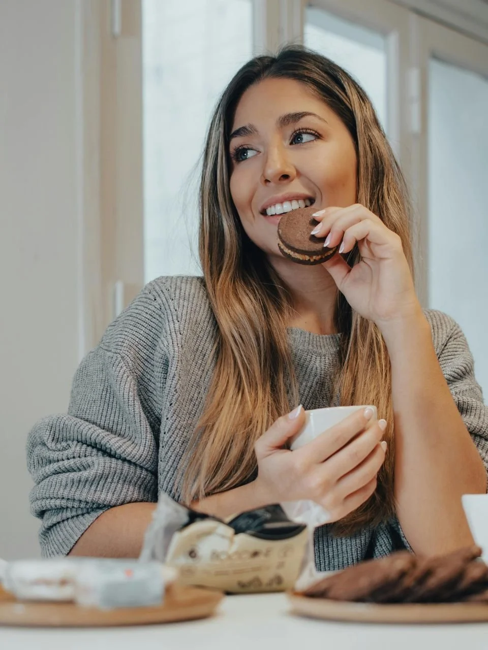 A woman with long brown hair and a gray sweater is enjoying a cookie and holding a cup, sitting at a table with food and snacks, near a window.