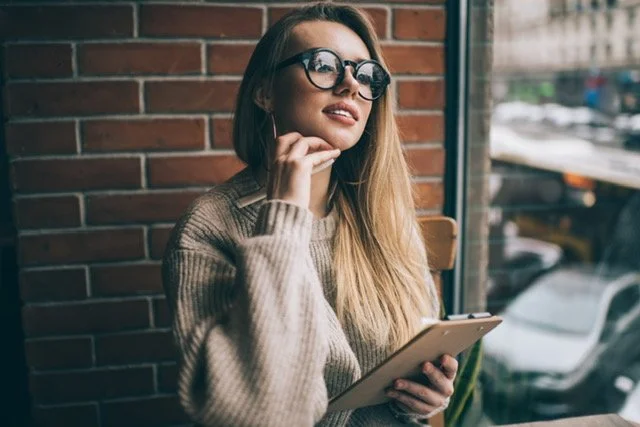 A young woman with long blonde hair wearing glasses and a beige sweater is sitting by a window in a cafe or restaurant, holding a tablet, with a brick wall behind her and cars visible outside.