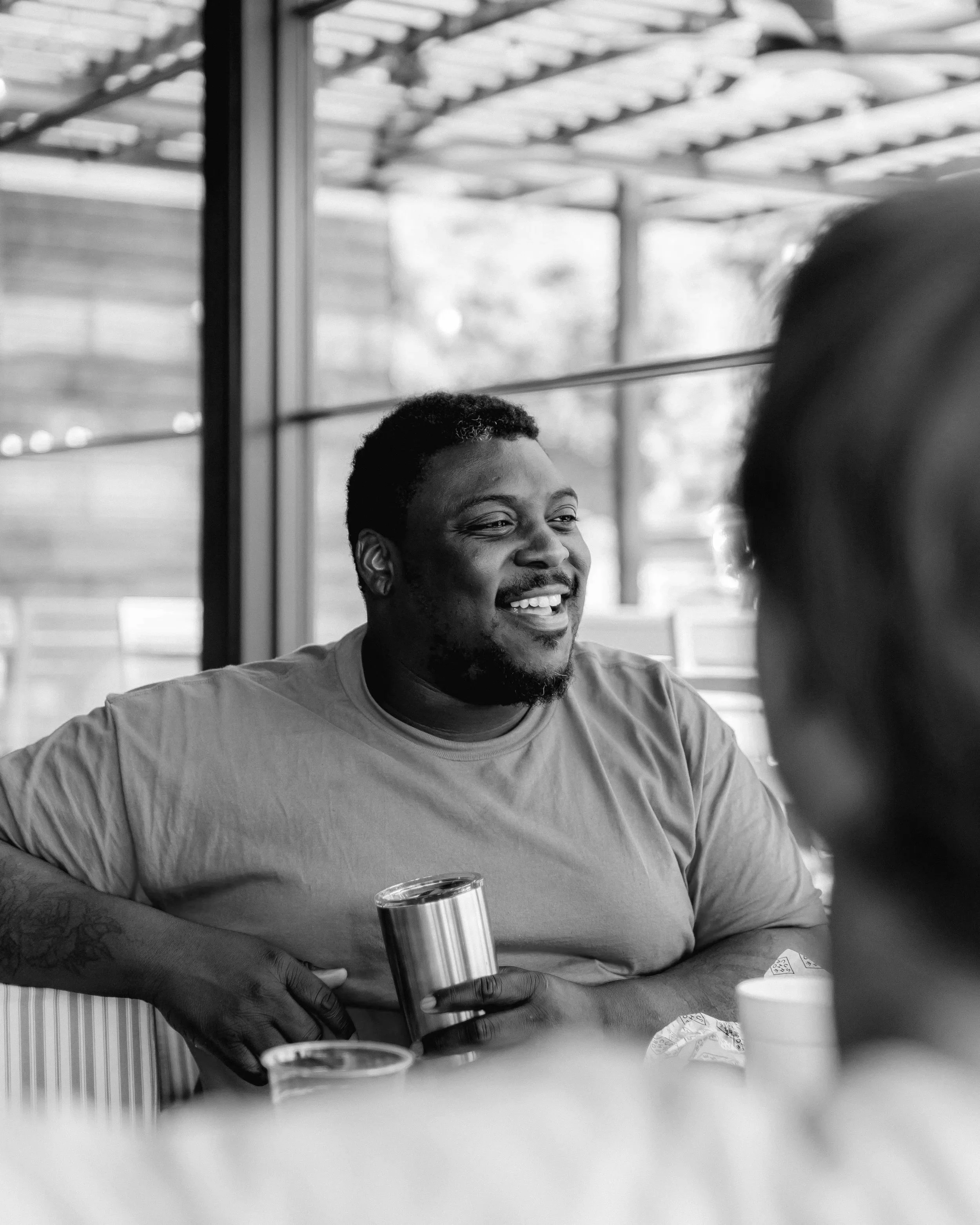 A man with short curly hair, a beard, and a big smile, sitting at a table with a beverage cup in his hand. He is wearing a light-colored t-shirt, and there are other cups and plates on the table. In the background, there are large windows and a wooden structure outside.