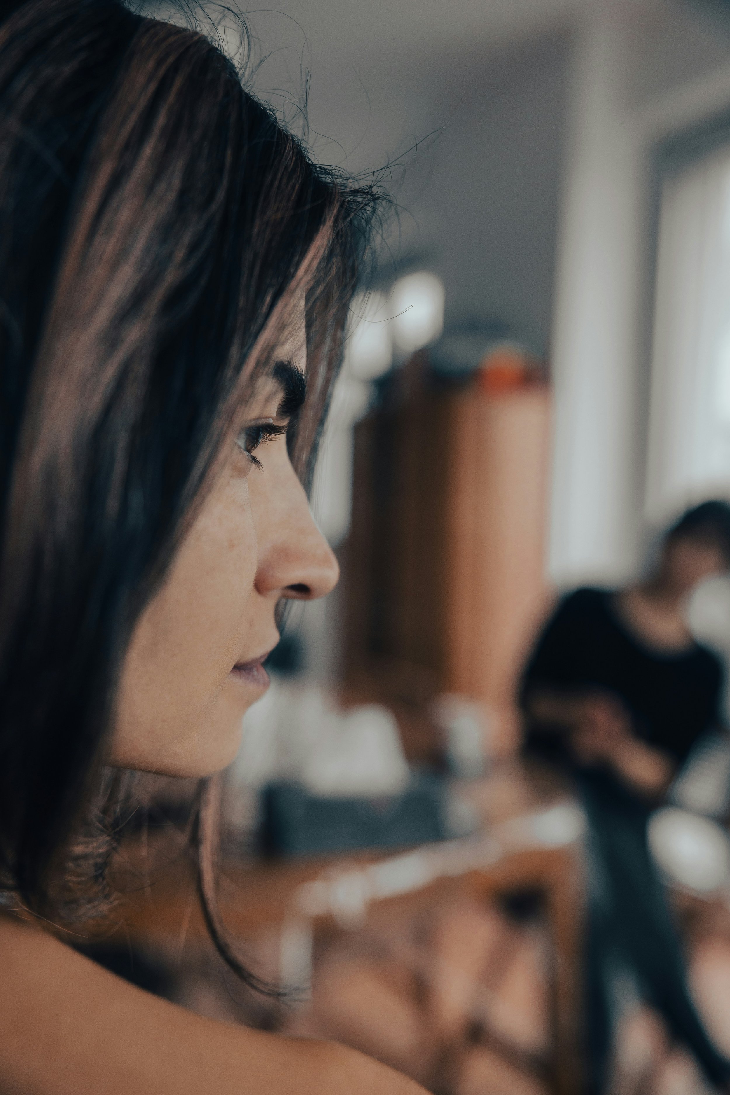 Close-up of a woman's face with dark hair, focusing on her eye and cheek, with blurred people working on computers in the background.