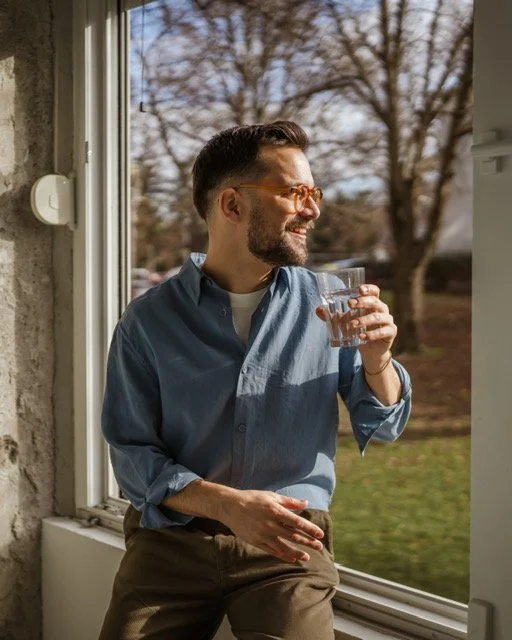 A man with glasses and a beard standing by a window, smiling, and holding a glass of water outdoors.