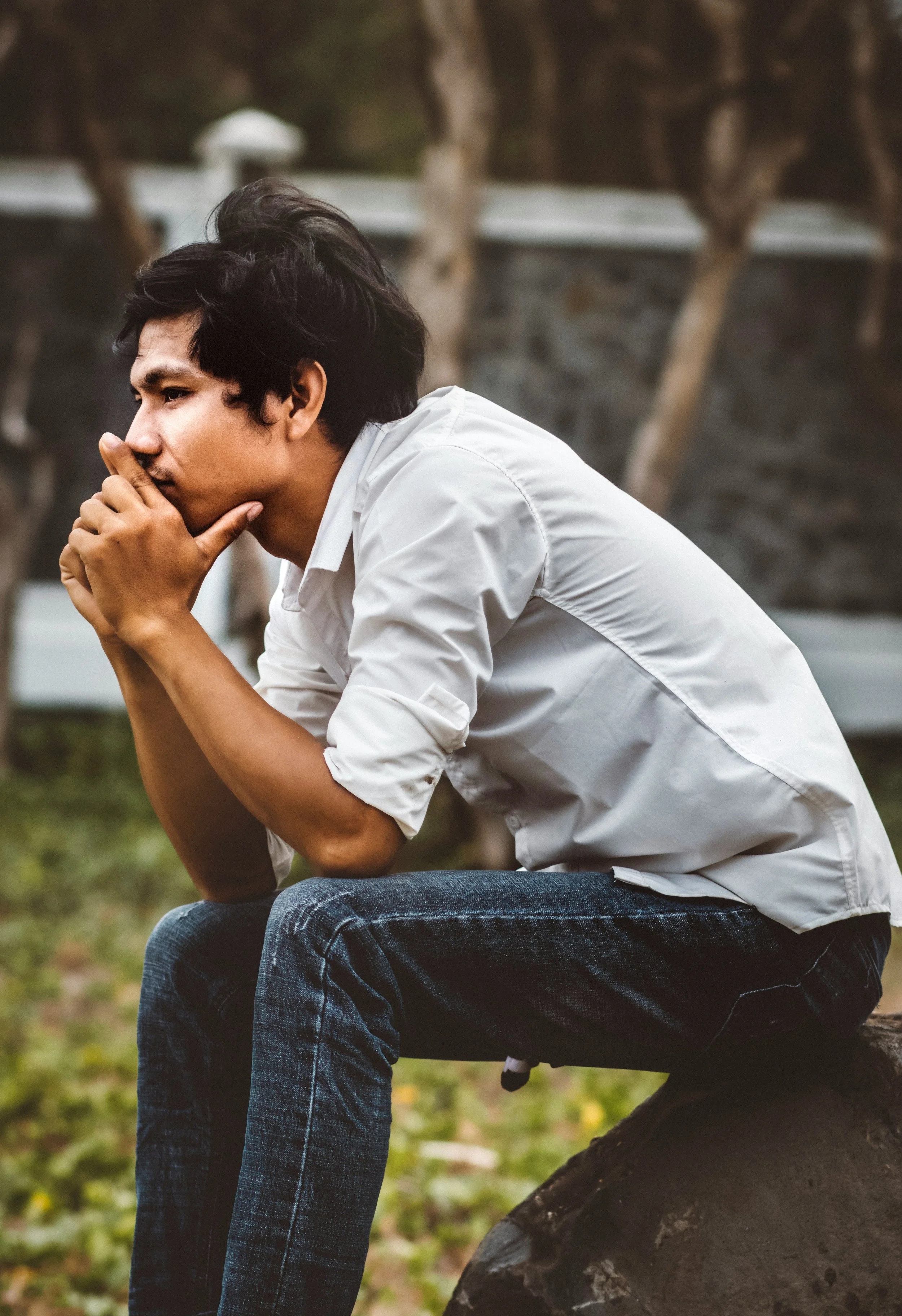 A young man with dark hair in a white shirt and jeans sitting on a log outdoors, resting his chin on his hand and looking thoughtfully into the distance, with trees blurred in the background.