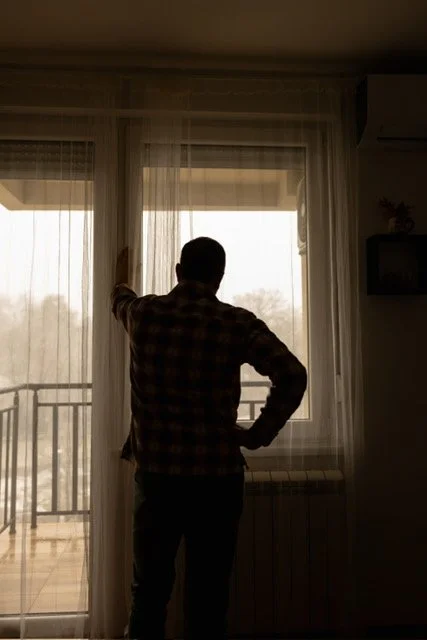 A person standing inside, looking out a large window with sheer curtains, with one hand on the curtain and the other on their hip.