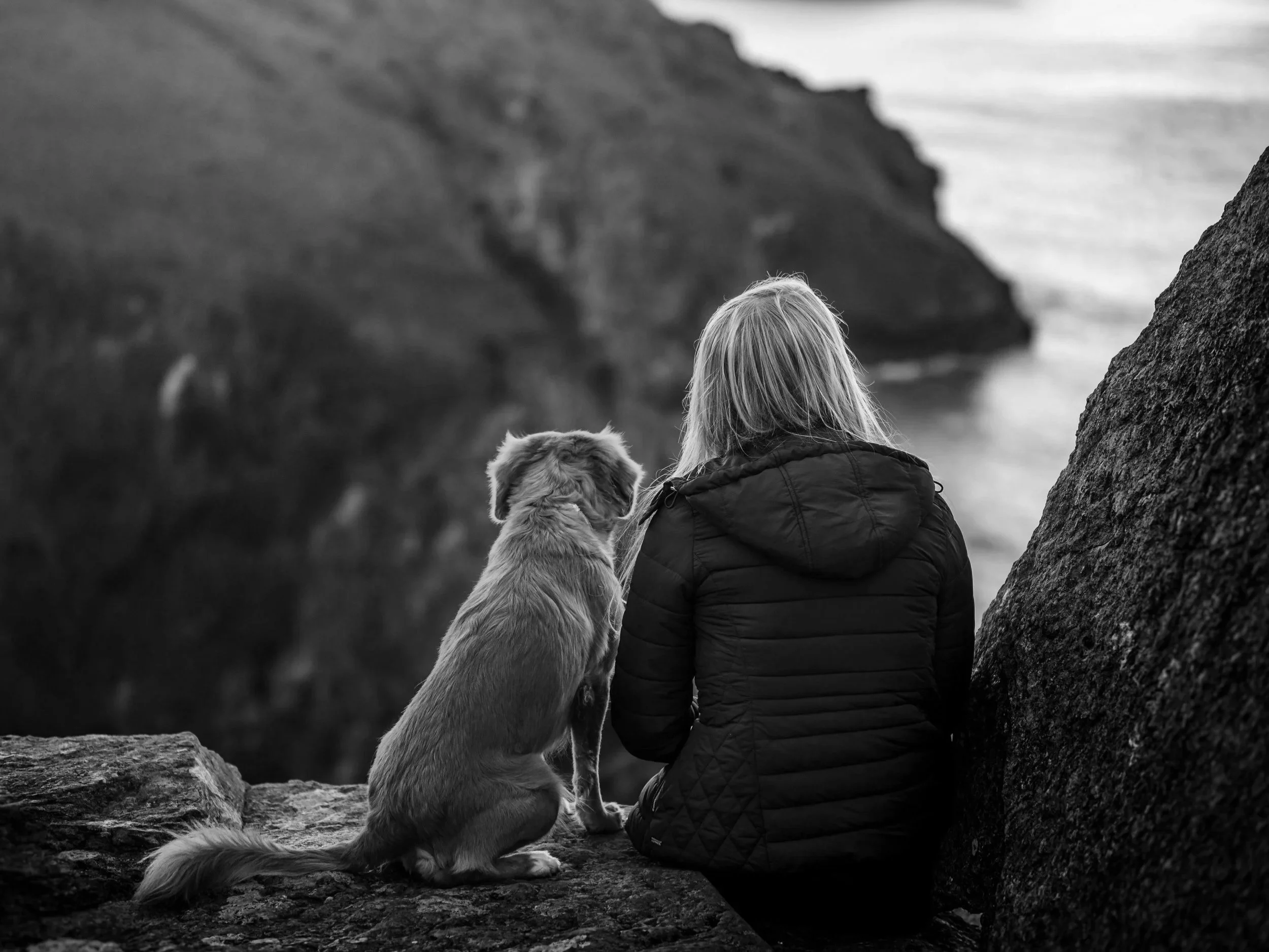 A person with long blond hair wearing a black puffer jacket is sitting on a rocky surface, near a large rock, looking out at a coastal landscape with cliffs and the ocean. The scene is in black and white.