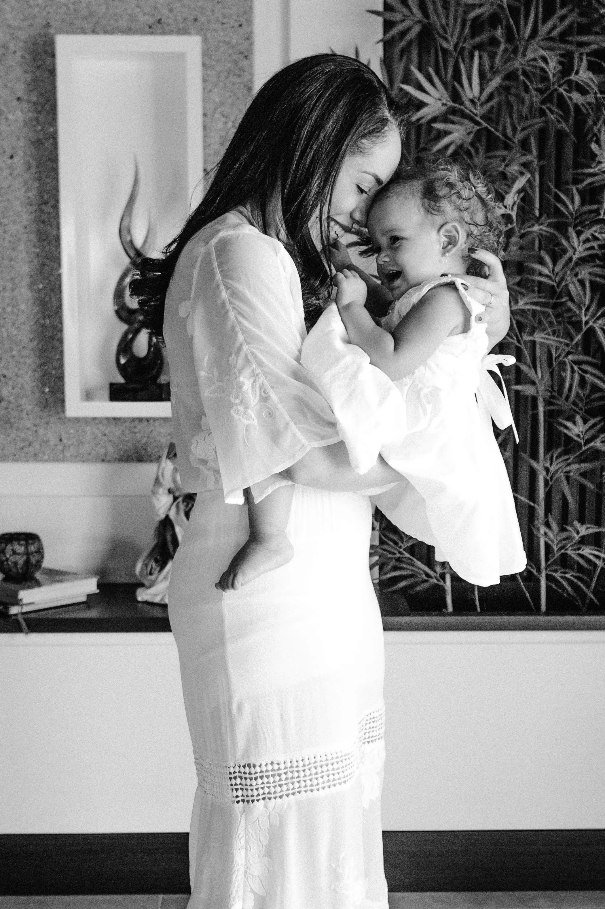 A woman in a white dress holding a young girl with curly hair, smiling and touching foreheads.