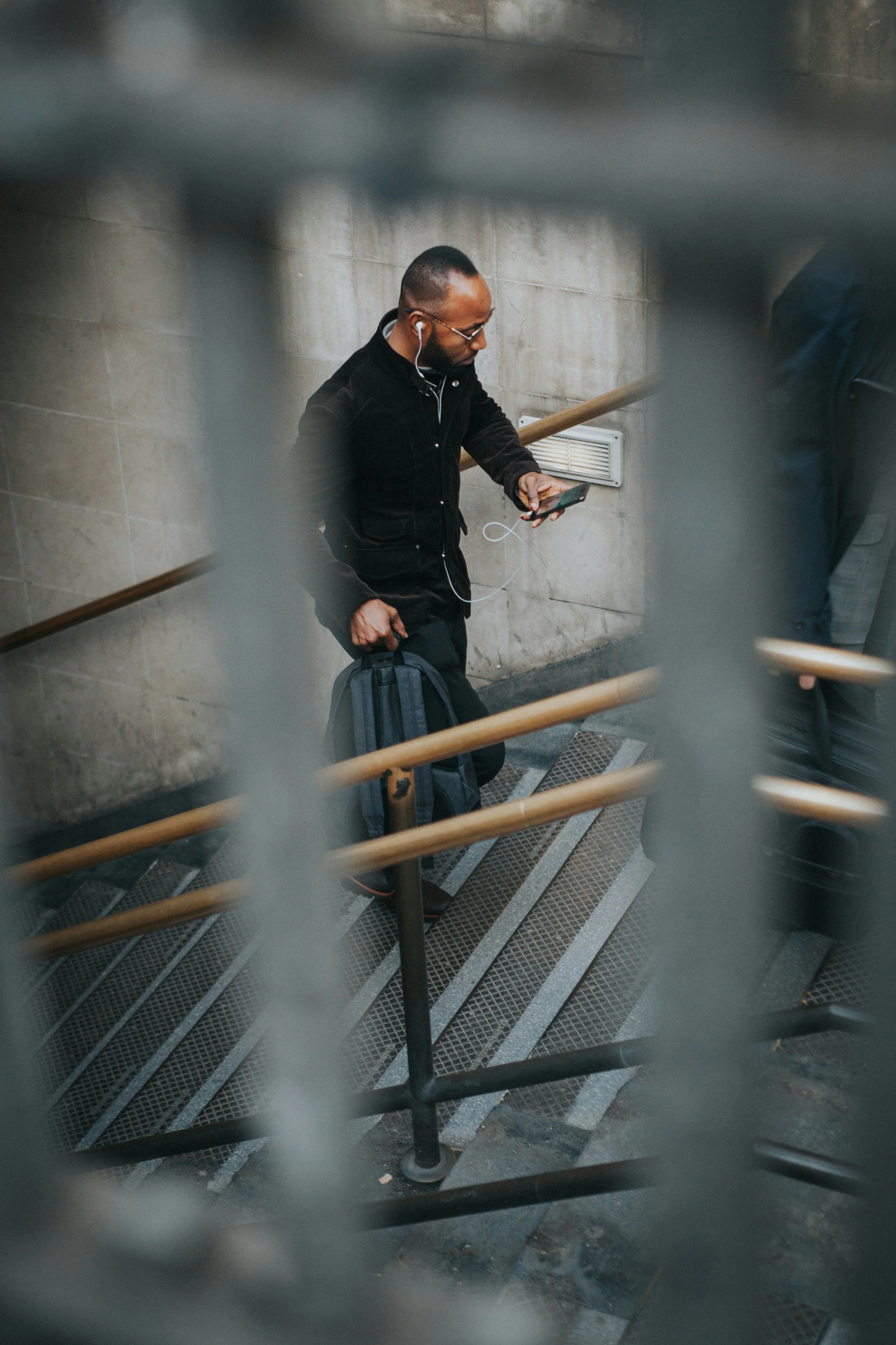 A man with glasses and a beard, wearing a black jacket, is looking at his smartphone while listening to earphones. He is holding a backpack in his left hand, standing on a stairway with metal steps and a wooden handrail, against a gray tiled wall.