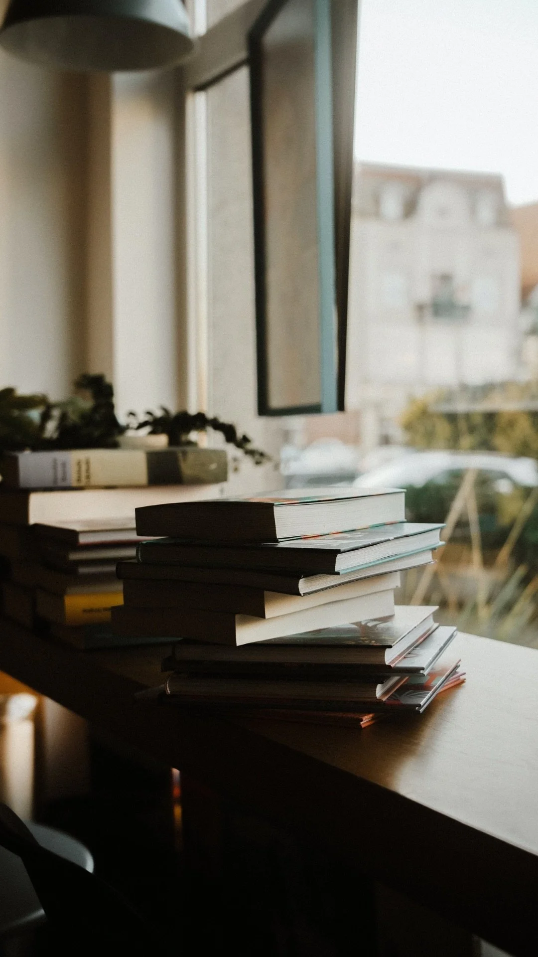 A window with a stack of books or magazines on a wooden surface indoors, with a blurred background of outside buildings and trees.