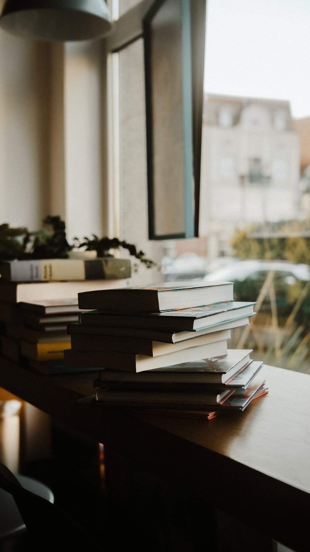 A window with a stack of books or magazines on a wooden surface indoors, with a blurred background of outside buildings and trees.