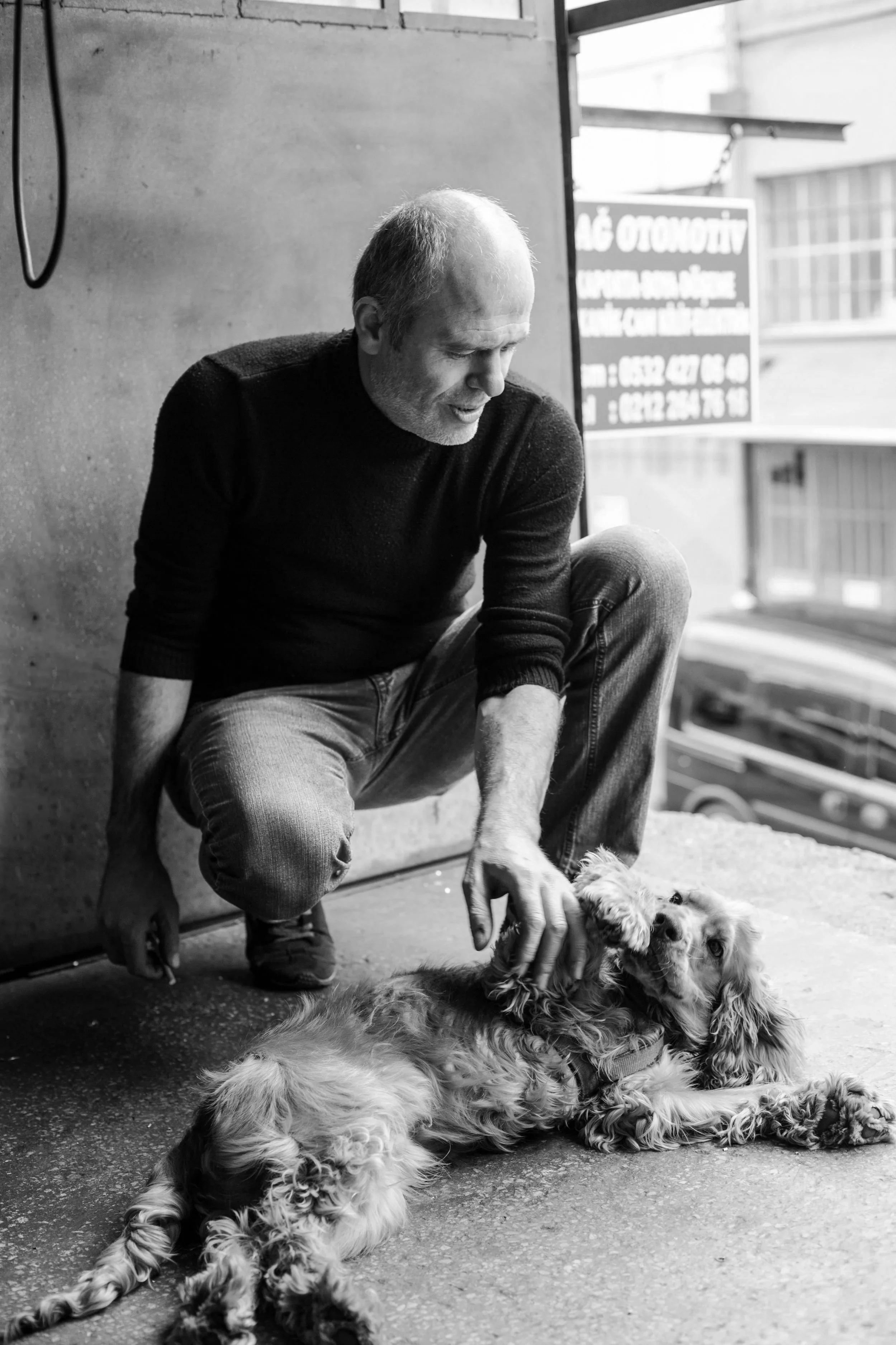 A man with short hair and a beard, dressed in a dark sweater and jeans, kneeling and petting a large, relaxed dog with long fur, lying on the ground, in a setting with a textured wall and a window with a sign and railings in the background.
