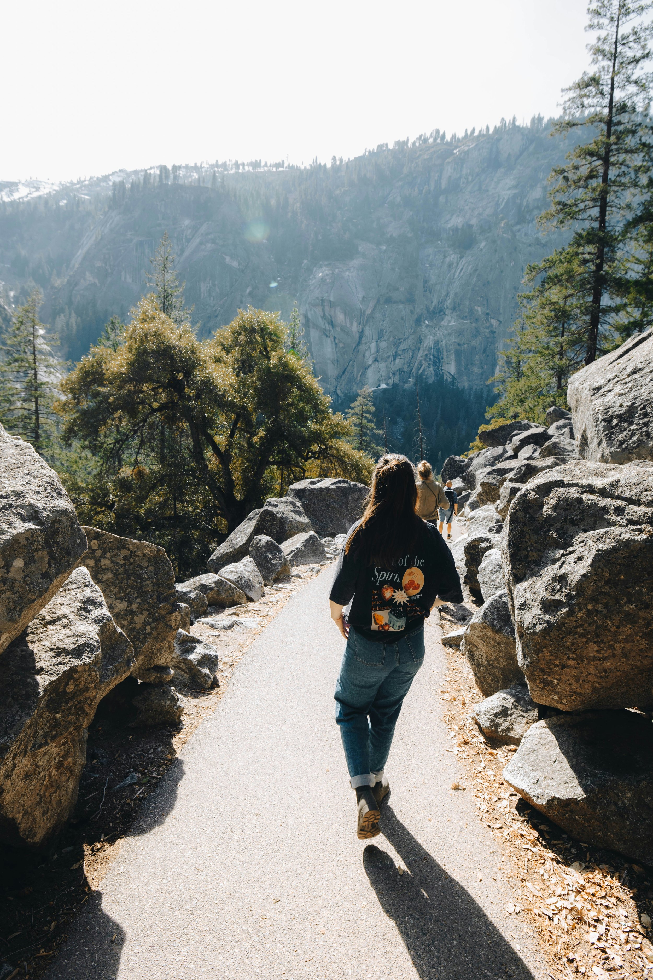 Person walking along a scenic mountain trail with large rocks on either side, tall pine trees, and mountains in the background under bright sunlight.