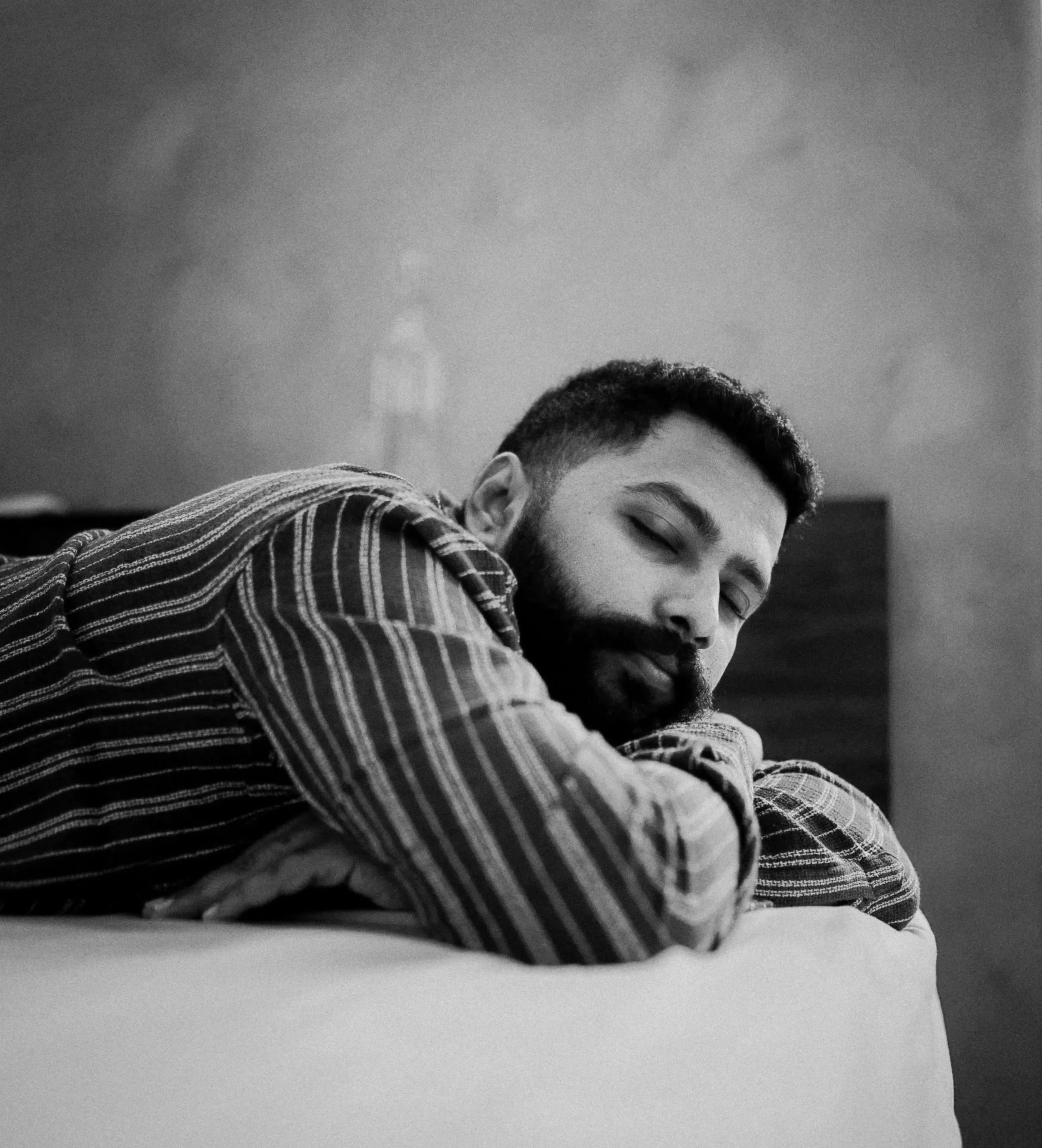 Black and white photo of a young man with a beard, closed eyes, and short hair, wearing a striped shirt, resting his head on his arm on a flat surface.