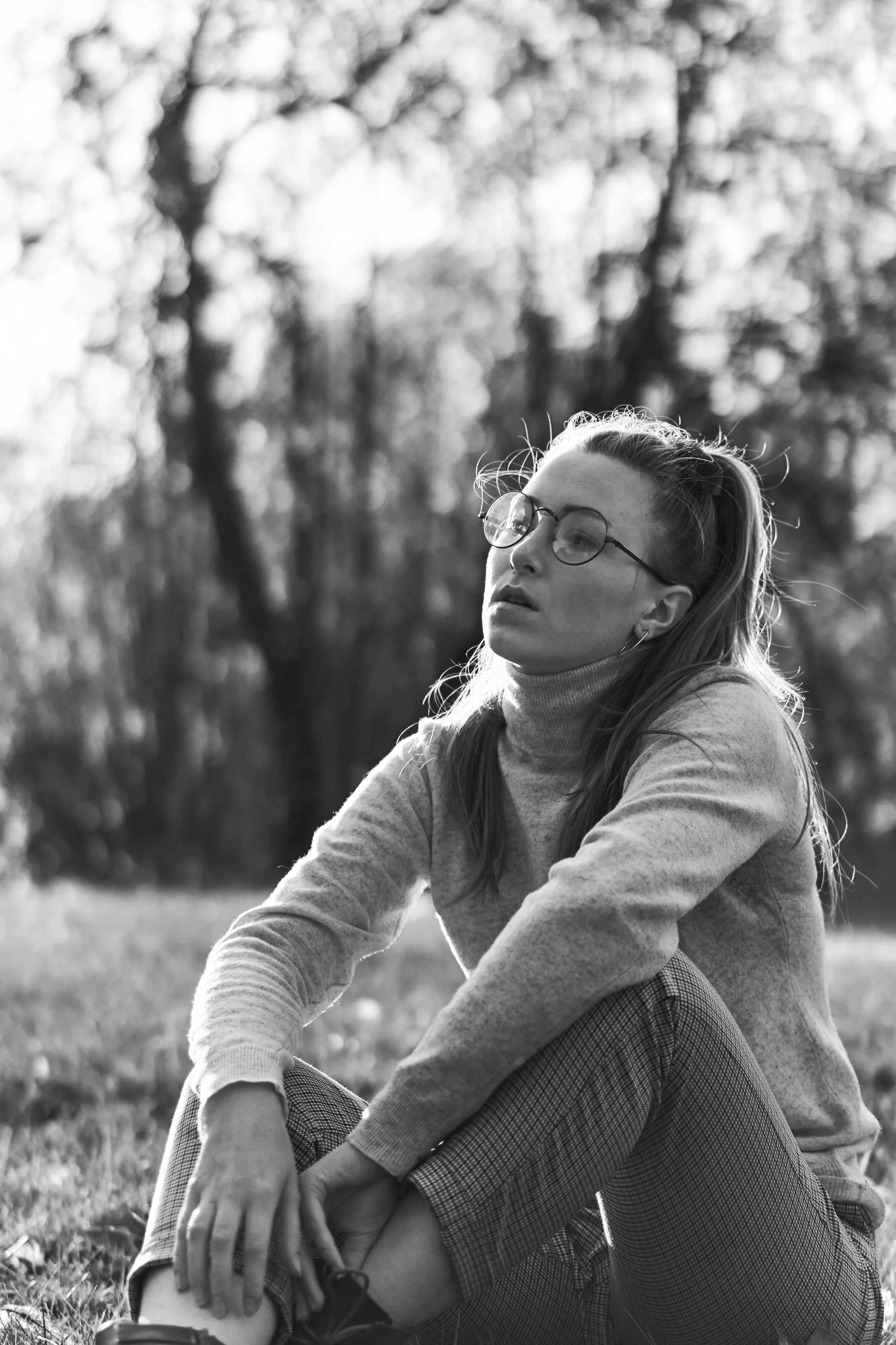 Black and white photo of a woman with glasses sitting outdoors on grass, with trees blurred in the background, looking off into the distance.