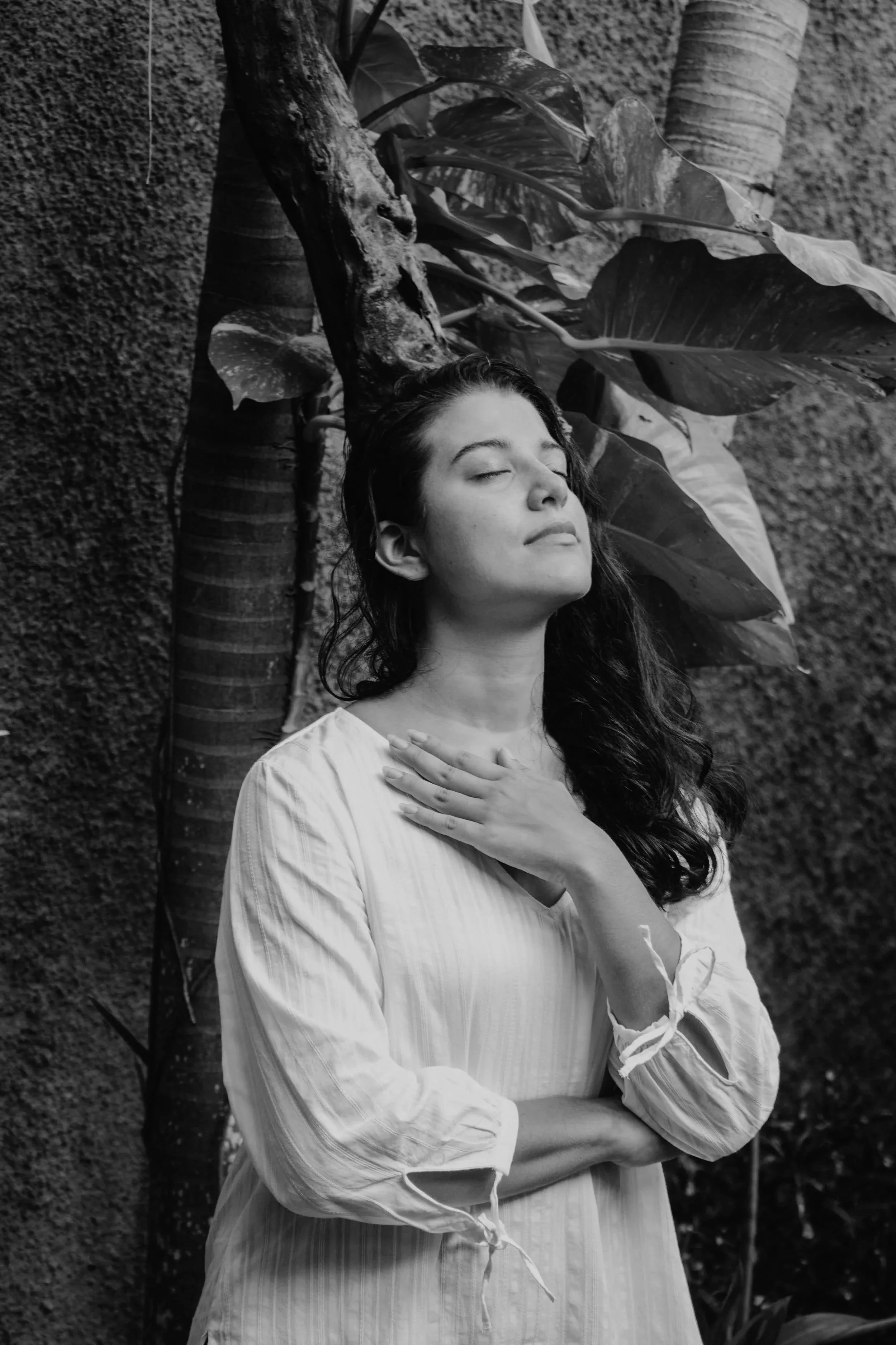 A woman with long dark hair and closed eyes, wearing a light-colored blouse, is standing outdoors in front of large broad leaves and a textured wall, with her hand over her chest.